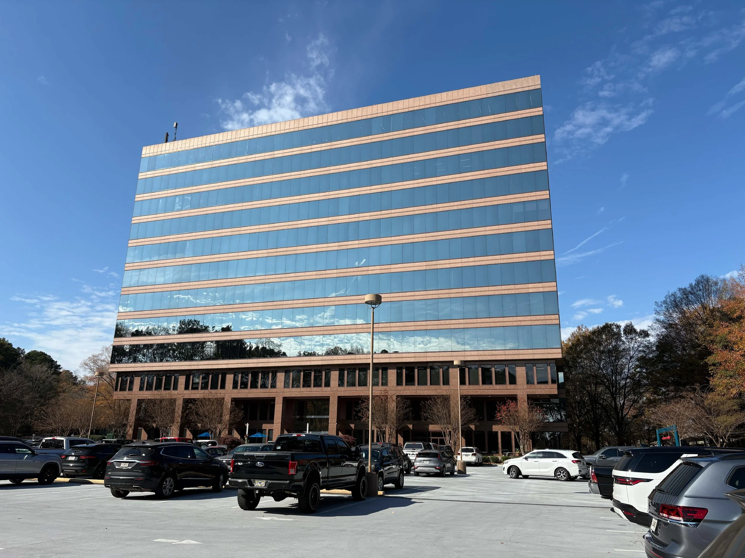 A modern office building with a reflective glass exterior, surrounded by a parking lot full of cars, trees, and a clear blue sky with some clouds.