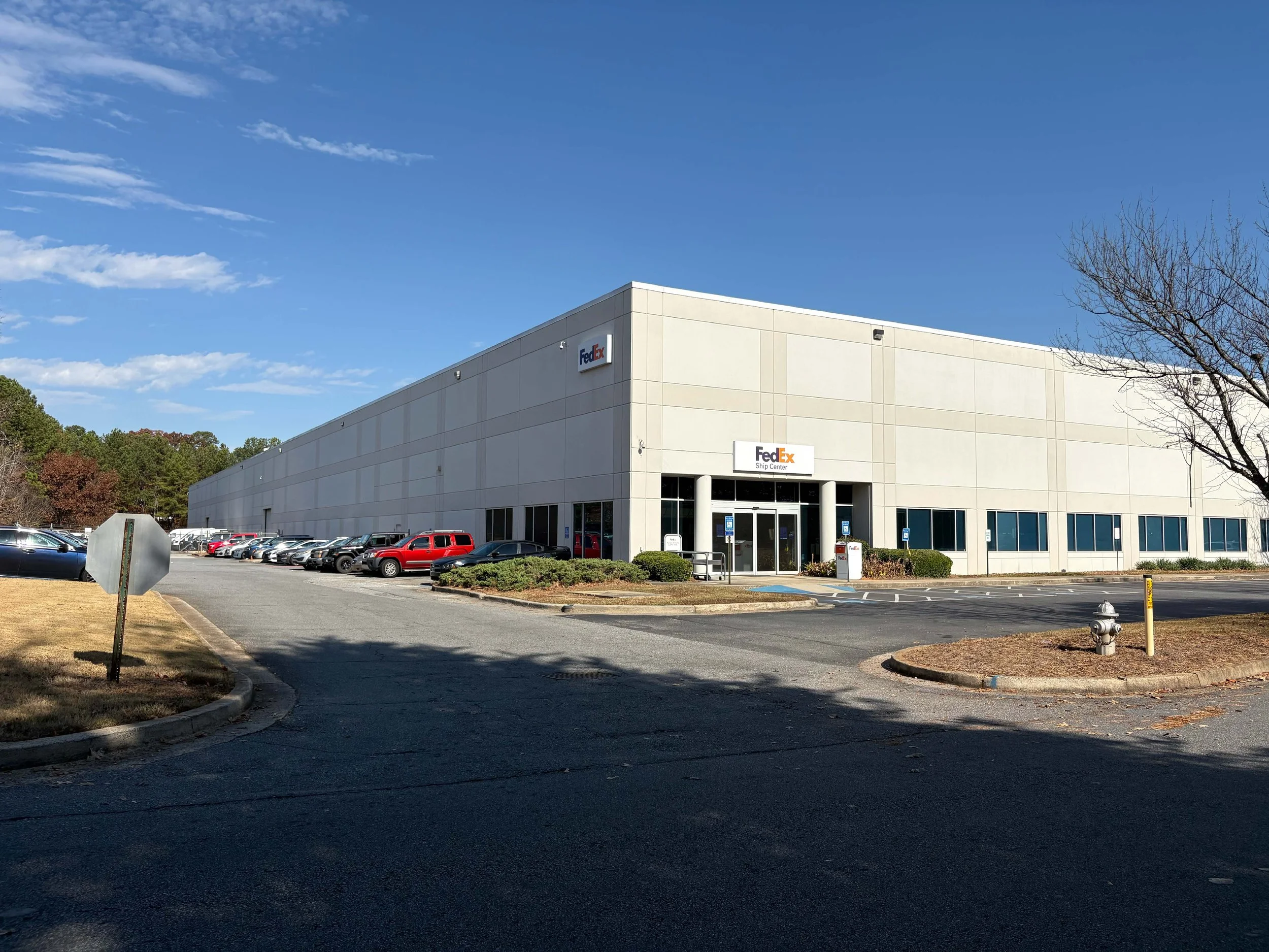 Exterior of a FedEx Ship Center with a large parking lot, several parked cars, some trees, and a clear blue sky.