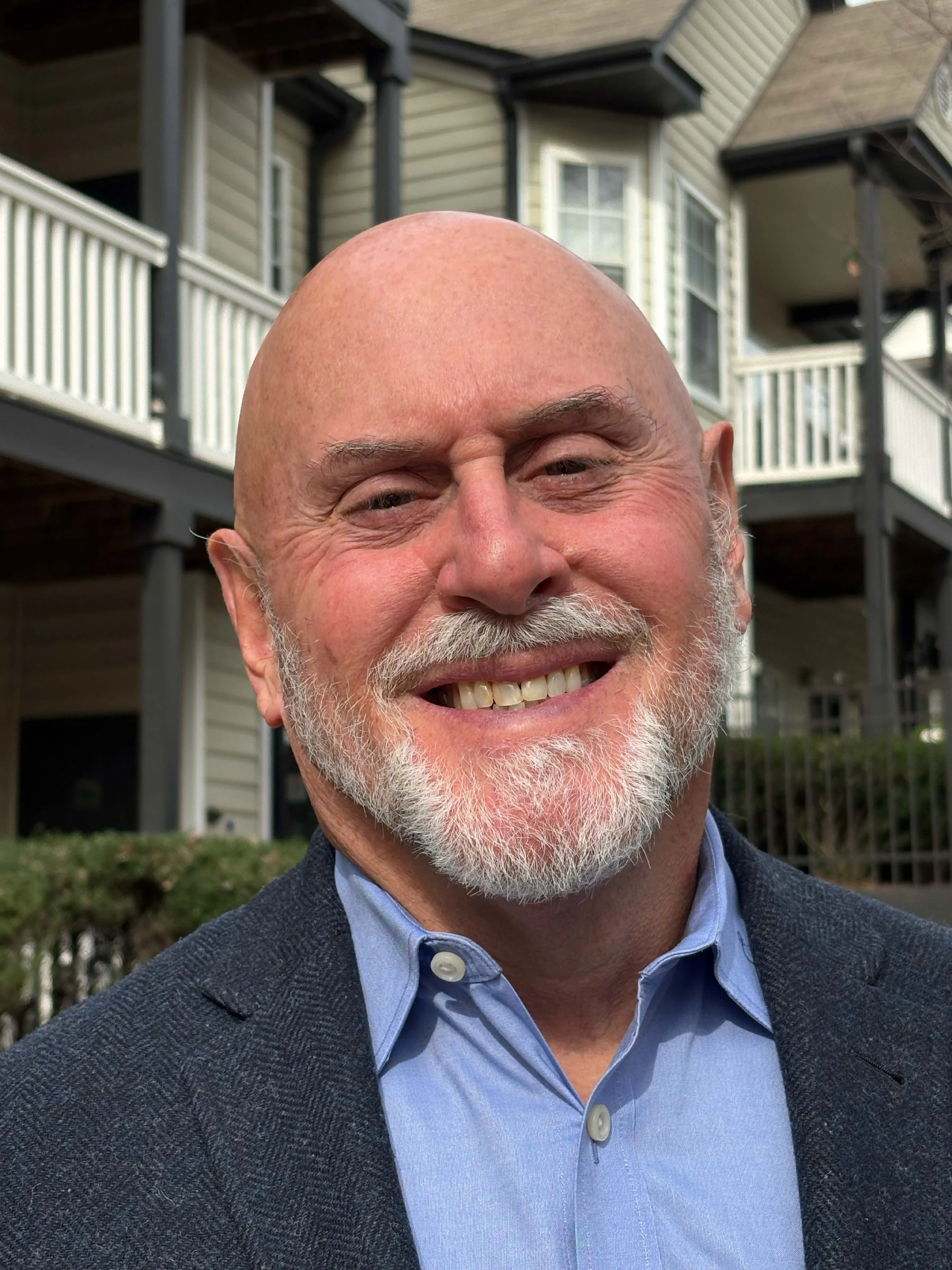 A smiling middle-aged man with a beard and bald head, wearing a blue collared shirt and a dark blazer, standing outdoors in front of a residential building with balconies and greenery.