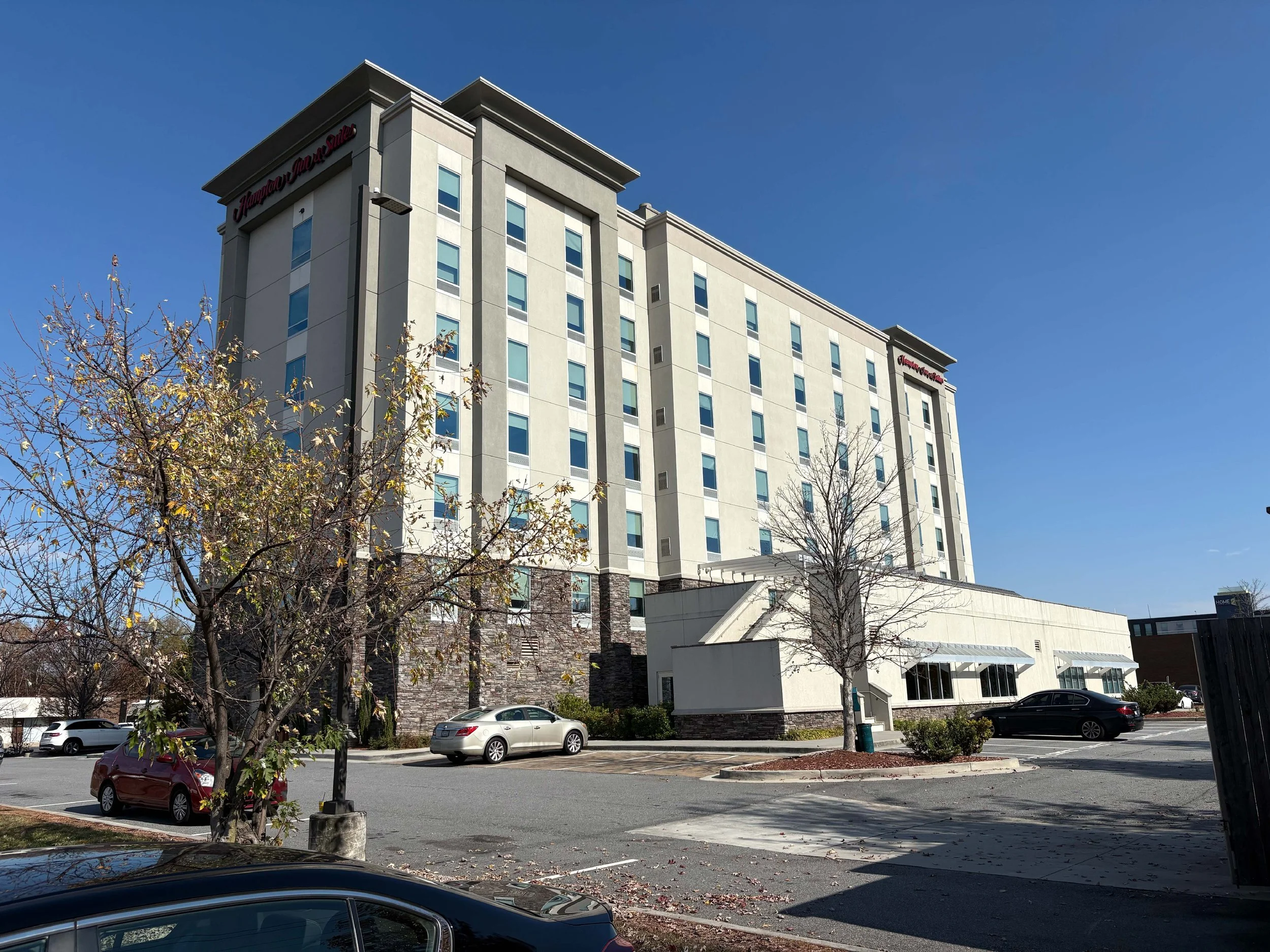 A tall hotel building with a sign reading 'Hampton Inn & Suites' at the top, surrounded by a parking lot with cars., blue sky, some trees with changing leaves, and a sidewalk in a suburban area.