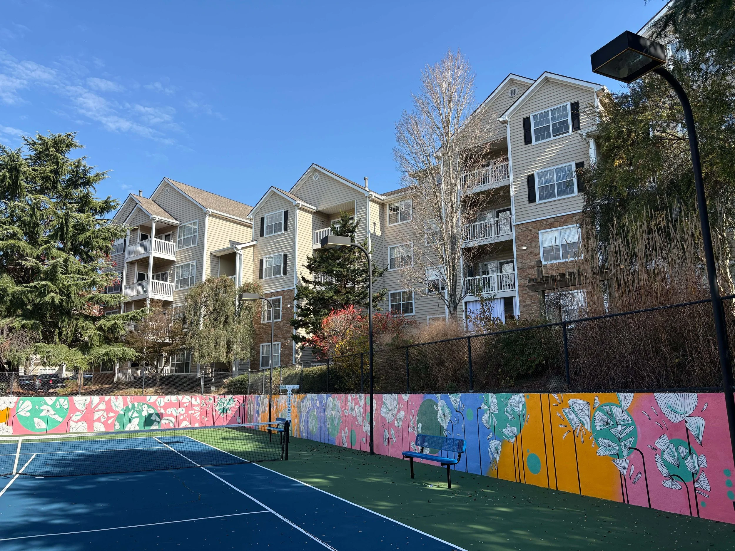 A tennis court with a colorful mural wall in the background, set against apartment buildings with balconies and trees, under a clear blue sky.