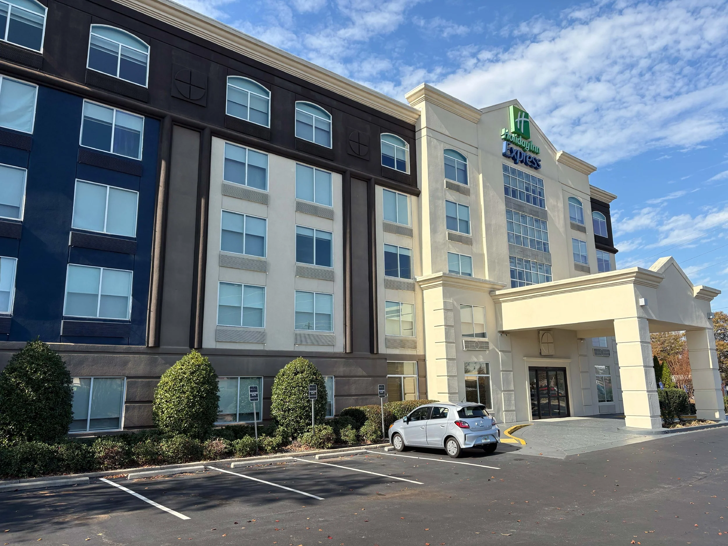 A Holiday Inn Express hotel building with parking spaces in front, a white car parked near the entrance, and a partly cloudy sky.