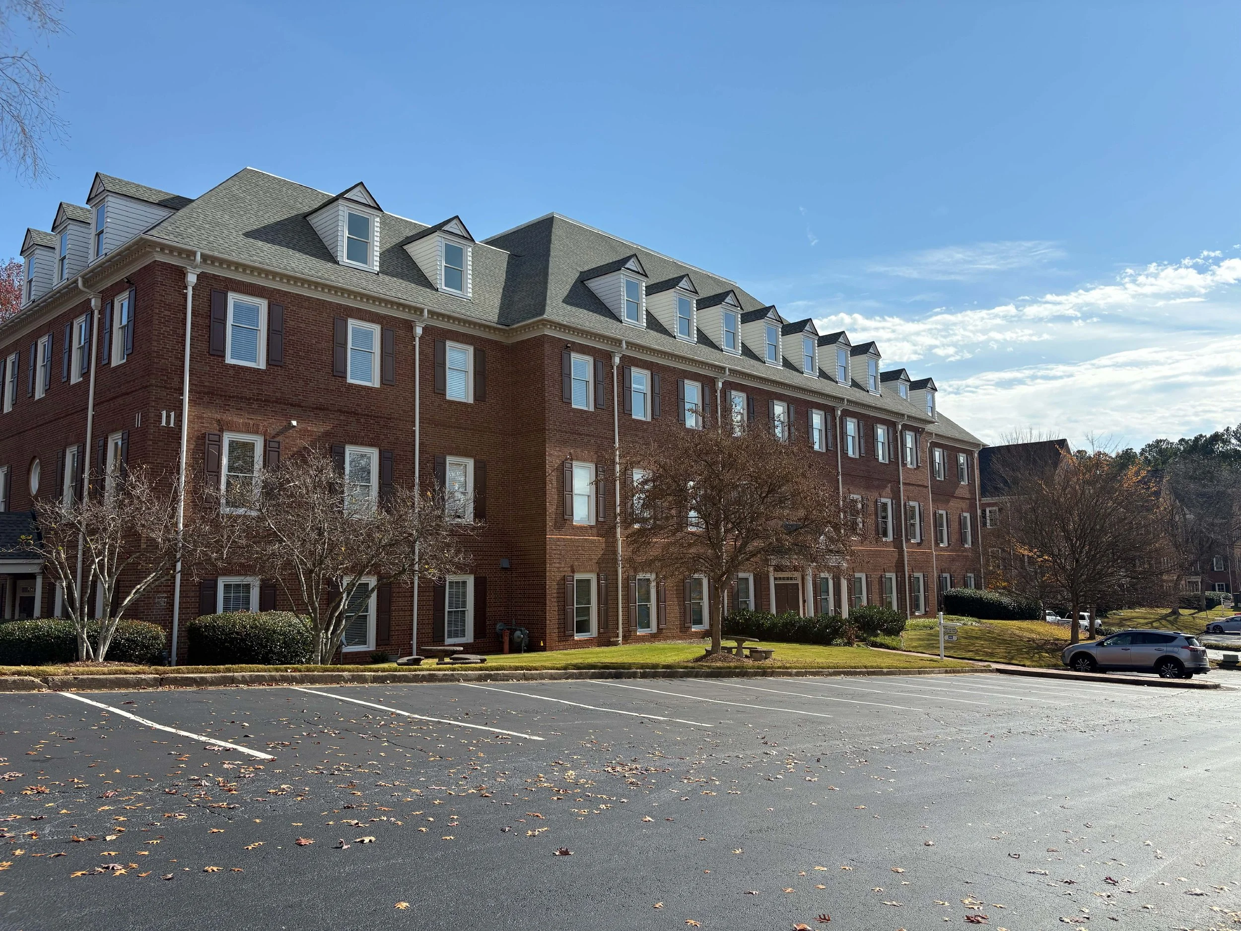 A multi-story brick apartment complex with gabled roofs, multiple windows with shutters, a parking lot in front, leafless trees, and a clear blue sky.