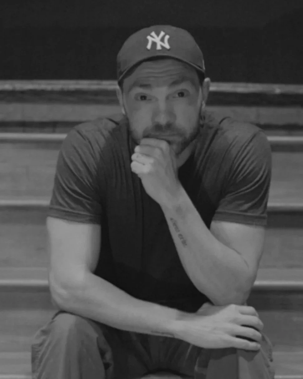 A young man wearing a New York Yankees cap and a dark t-shirt, sitting on bleachers with his chin resting on his hand, looking at the camera.