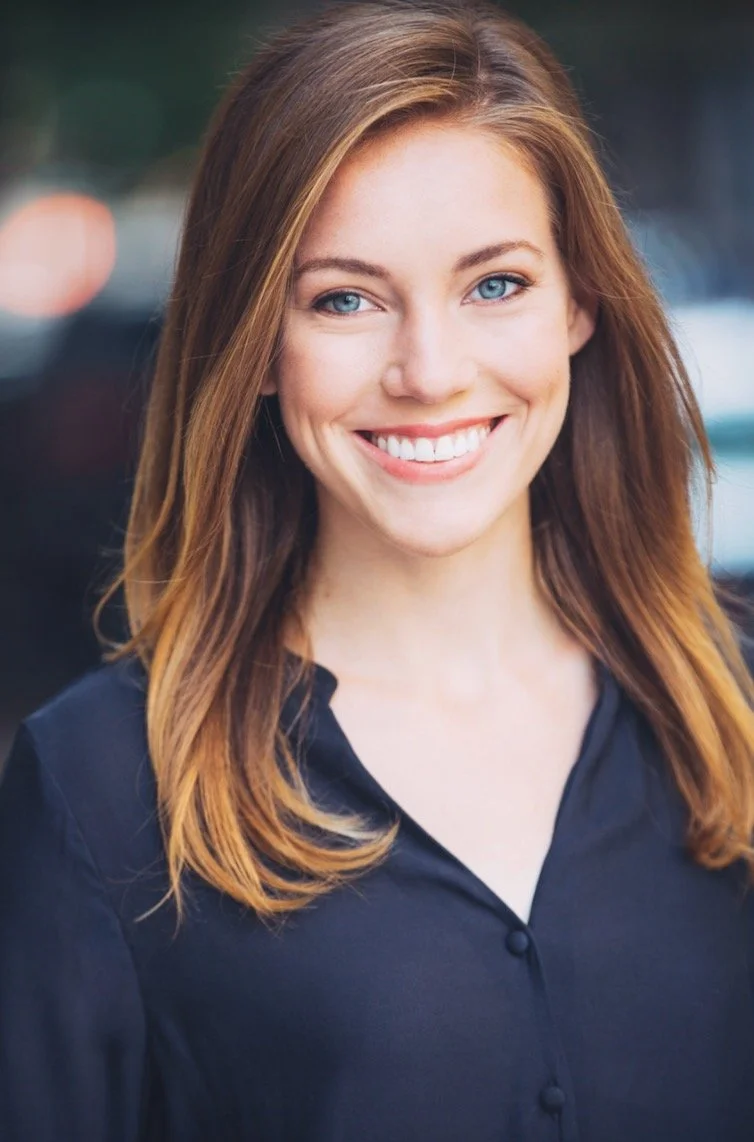 A young woman with long, wavy red hair and blue eyes smiling outdoors, wearing a black top.