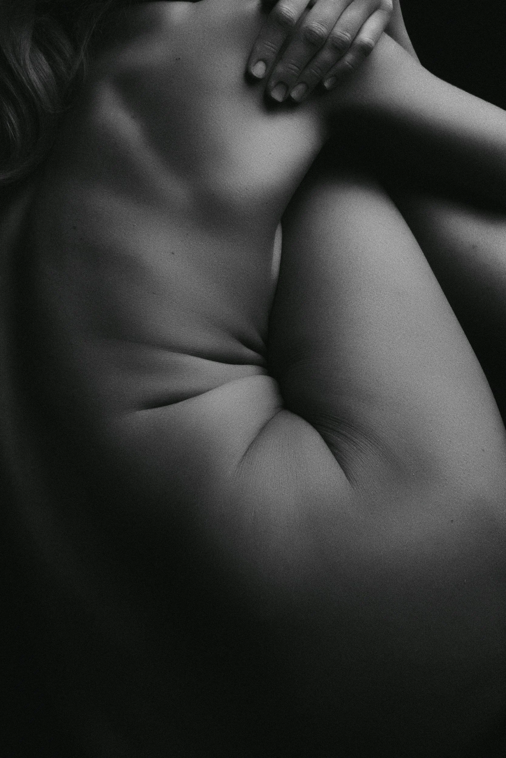 Black and white close-up photograph of a person's upper body, with one hand resting on their chest and their head turned away. The focus is on the contours of the shoulder, arm, and neck with a textured background.