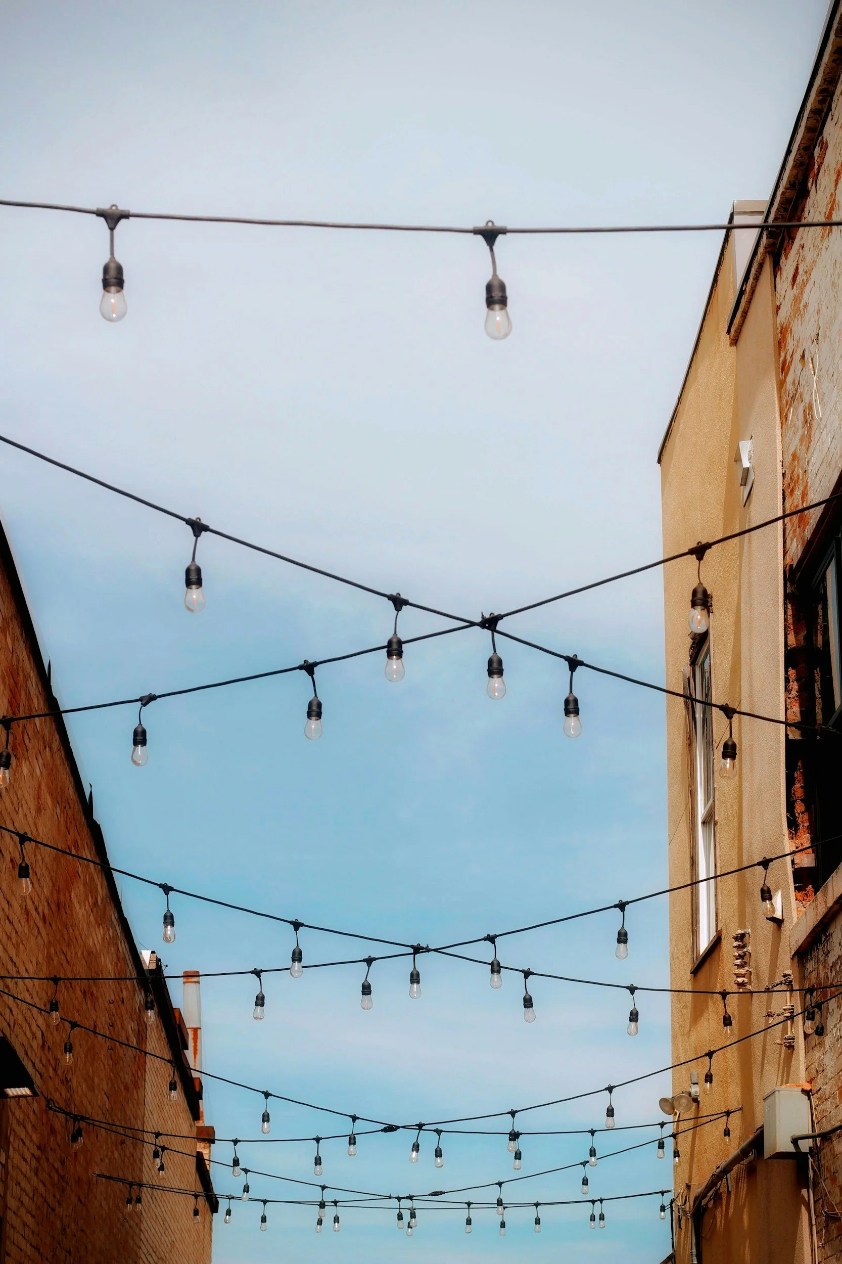 String of hanging outdoor string lights decorated between brick and stucco buildings, against a cloudy sky.