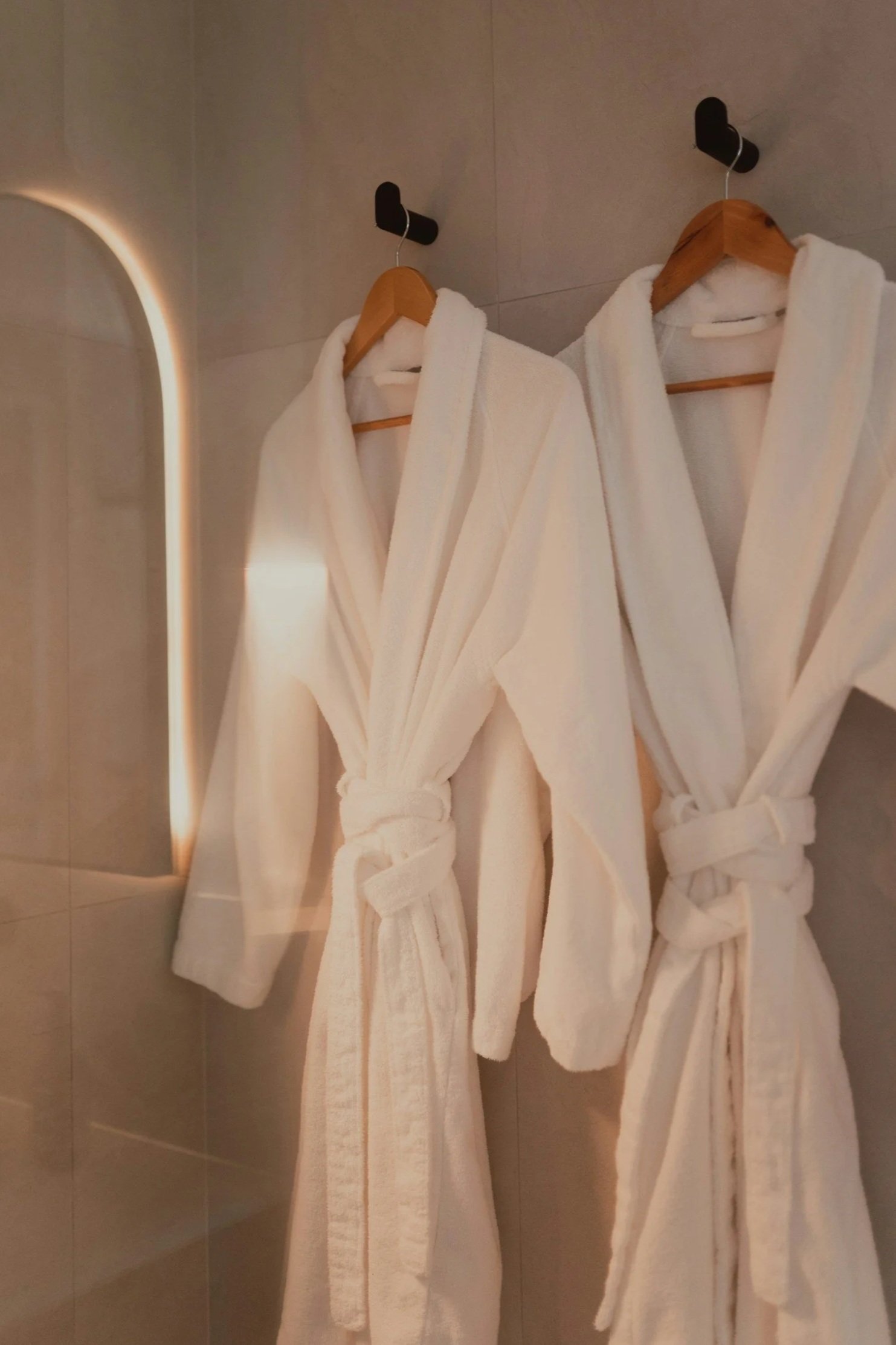Two white bathrobes hanging on wooden hangers on black hooks on a tiled wall, next to a mirror with a lighted border.