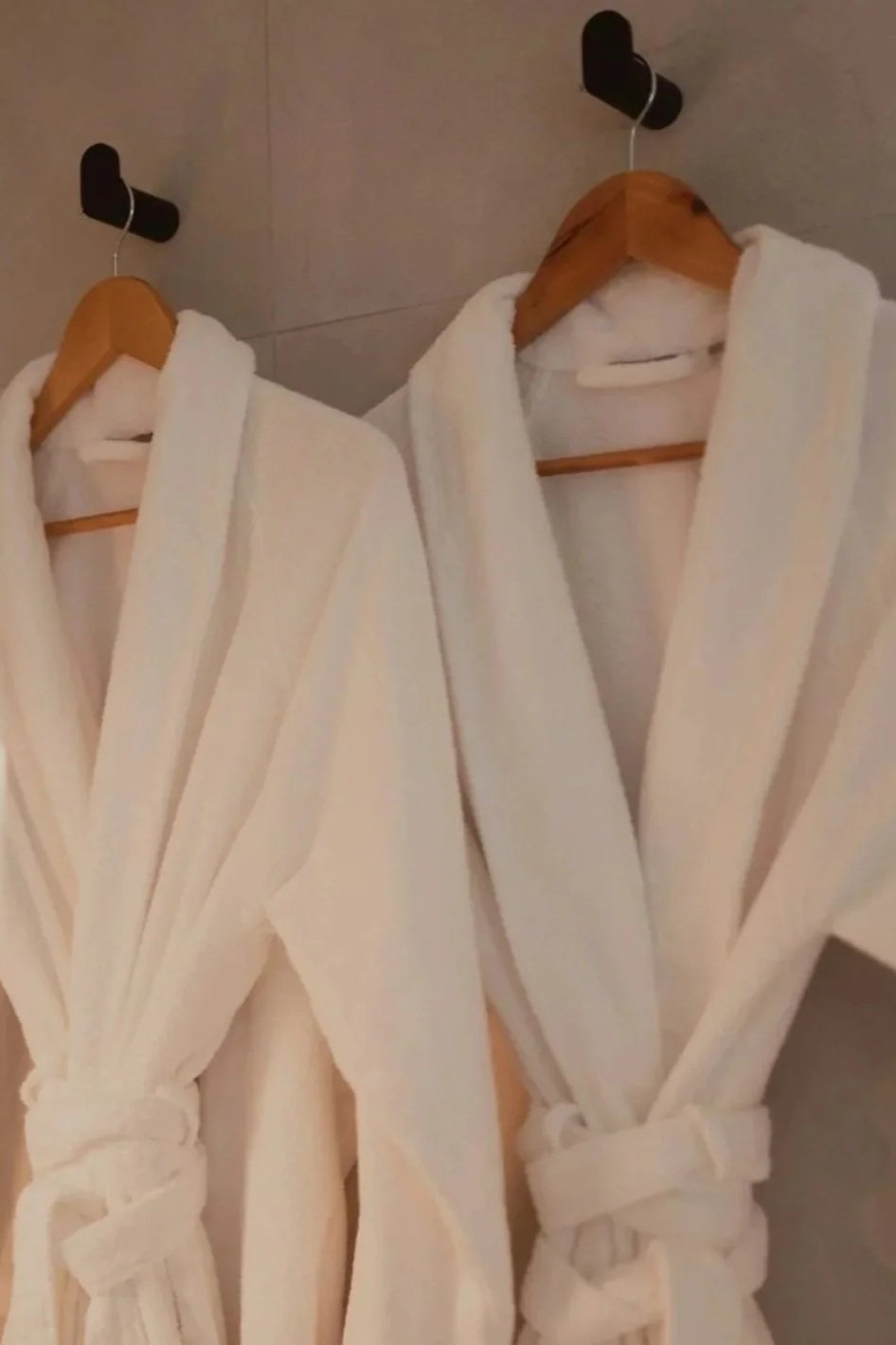 Two white bathrobes hanging on wooden hangers on black hooks on a tiled wall, next to a mirror with a lighted border.