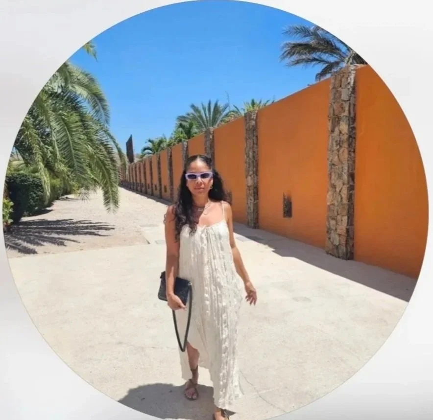 Woman in a white maxi dress walking near an orange wall with stone accents on a sunny day, wearing sunglasses and carrying a purse.