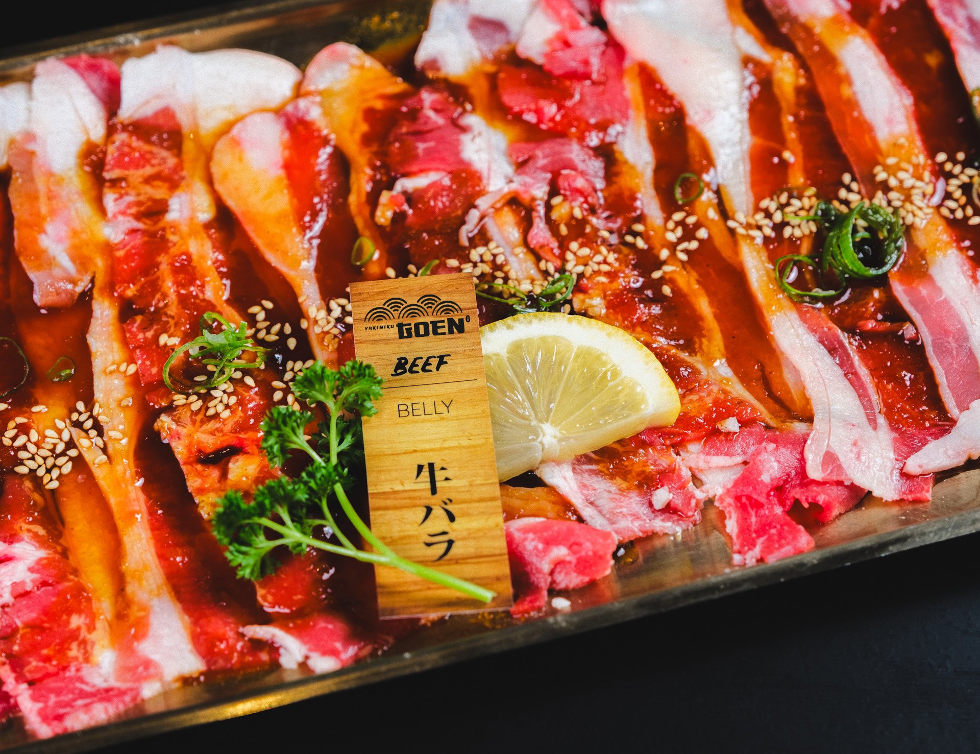 Thin slices of raw beef with sesame seeds, chopped green onions, and a lemon wedge on a tray, labeled 'TOEN BEEF BELLY' in Japanese and English.