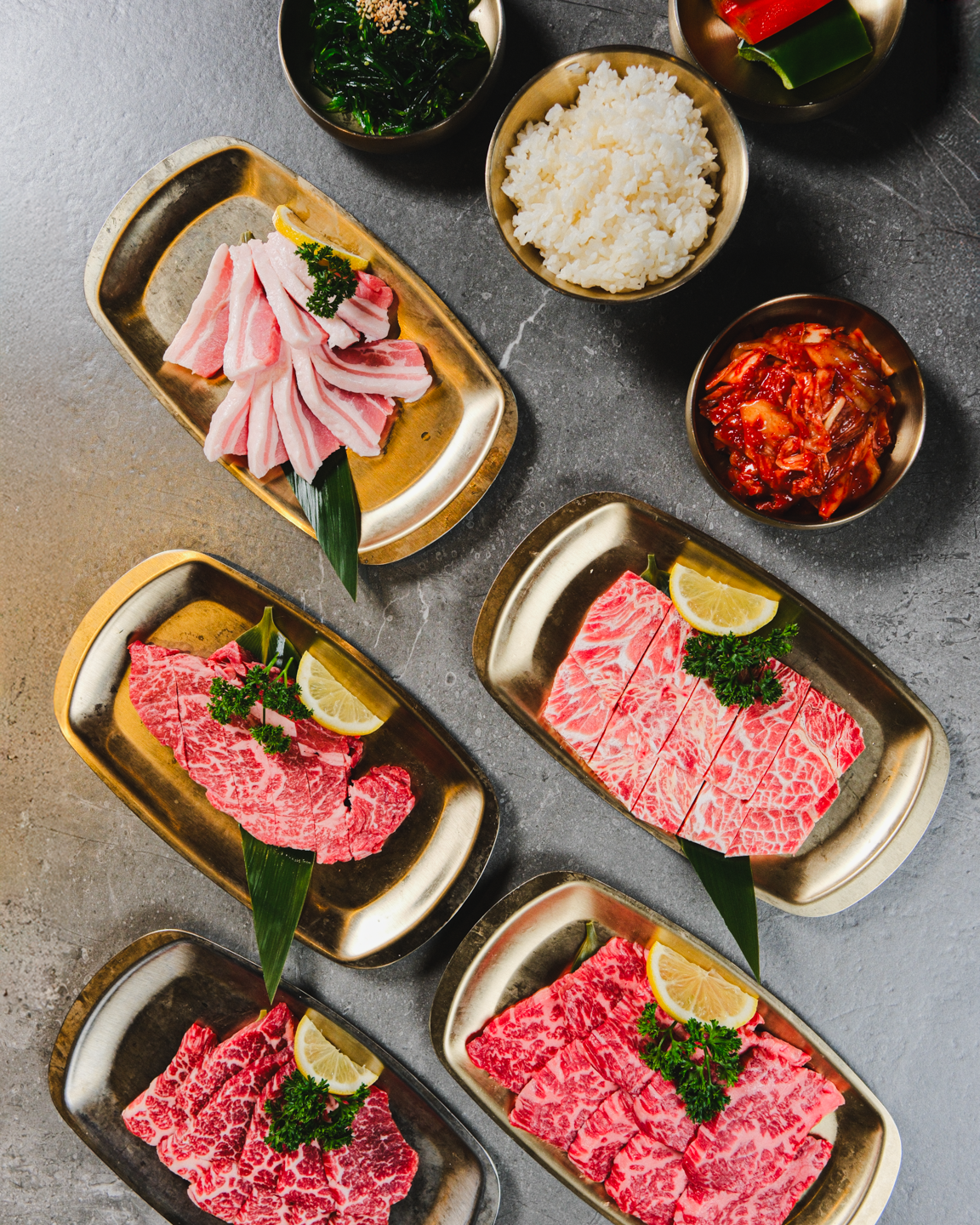 Assorted cuts of marbled raw beef and pork on gold trays, garnished with lemon slices and parsley, with side dishes of rice, kimchi, and vegetables on a gray surface.