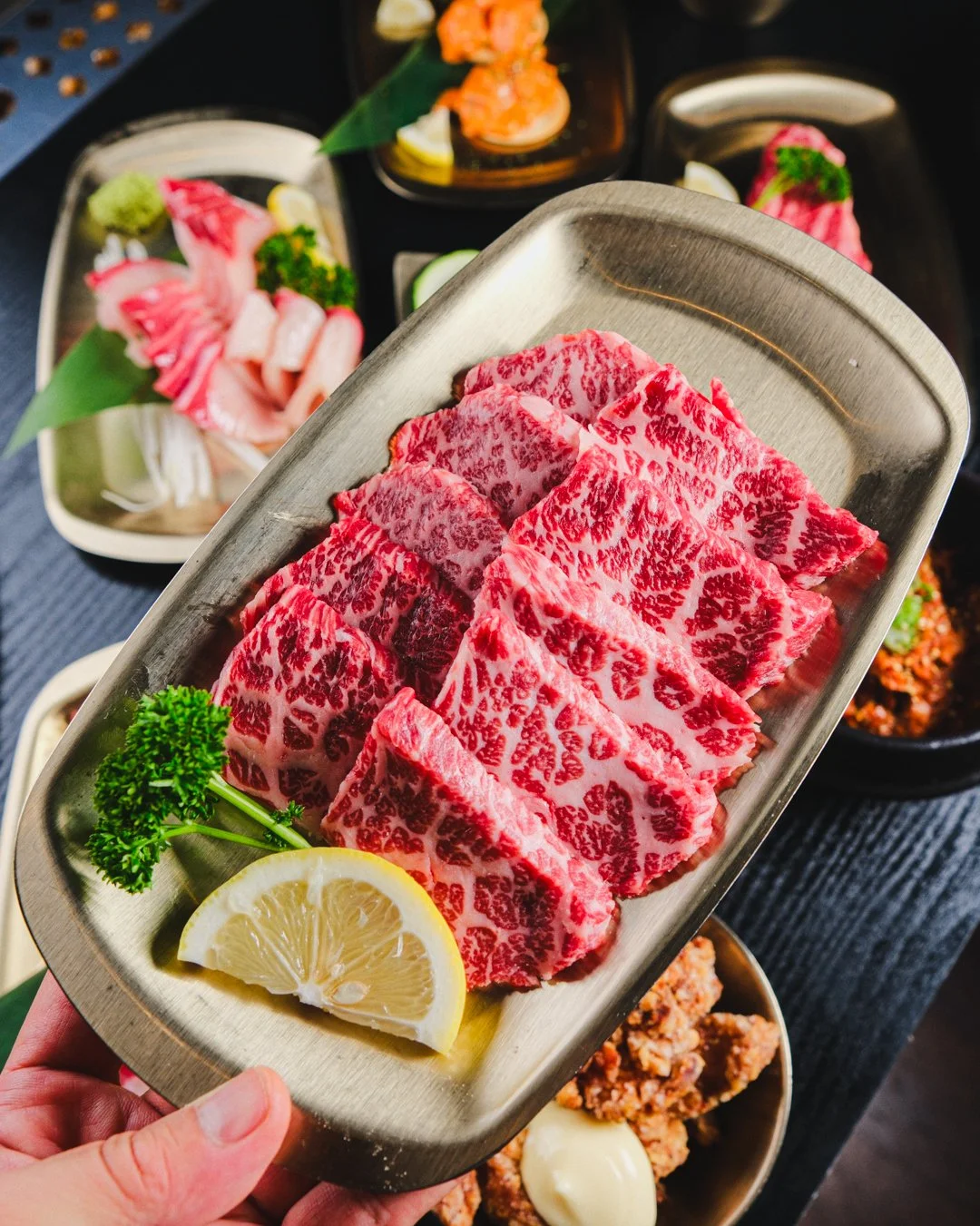 Slices of marbled Wagyu beef on a metal tray, garnished with lemon wedge and parsley, with assorted sashimi and fried foods in the background.