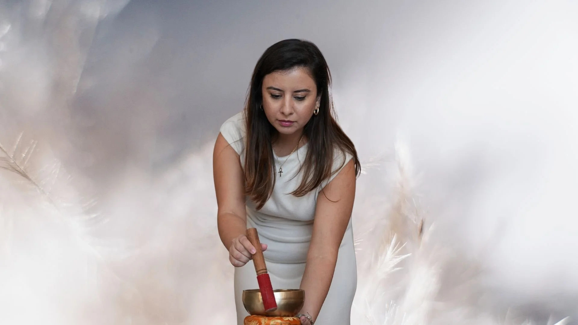 A woman in a white dress holding a singing bowl and a wooden mallet, with a cloudy, feather-like background.