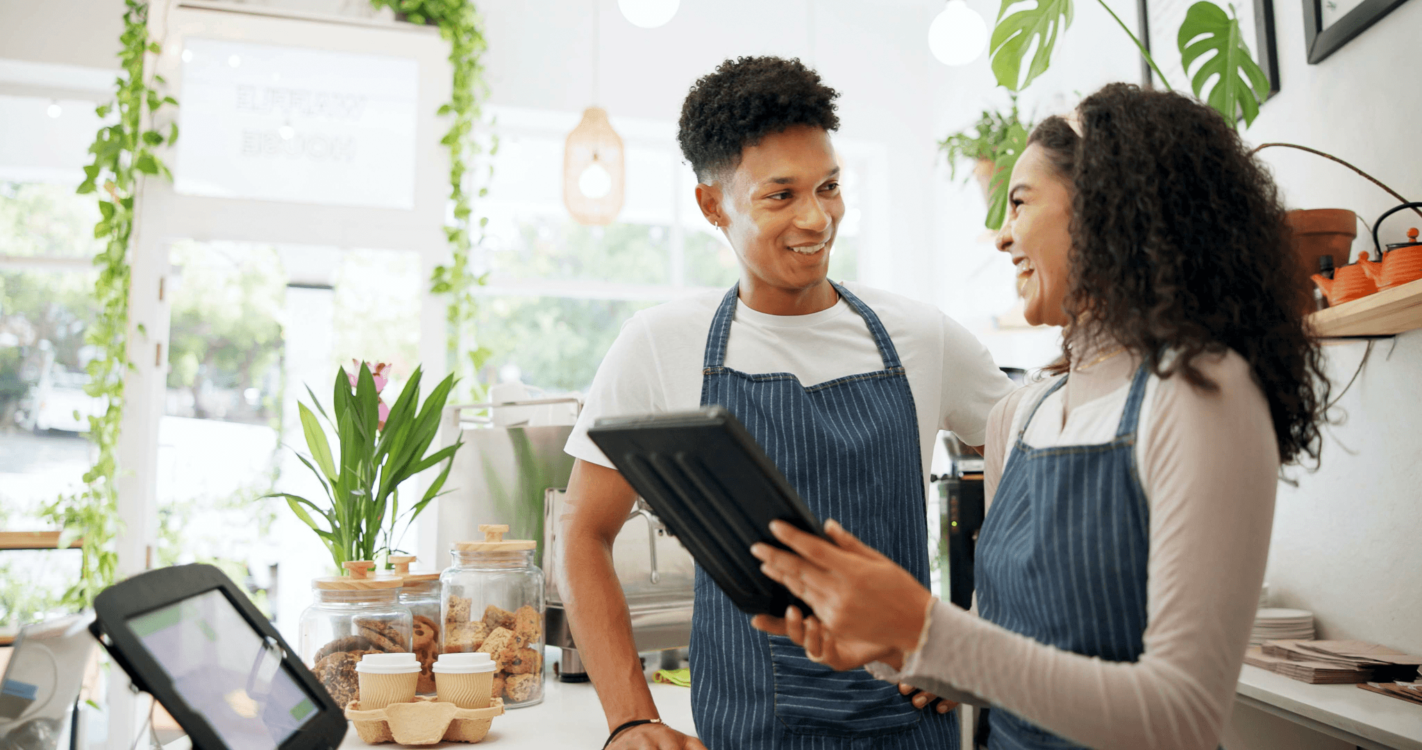 Two smiling baristas in aprons working behind the counter of a bright, modern coffee shop. One is holding a tablet while discussing with her colleague. The shop is decorated with green plants and jars of cookies.