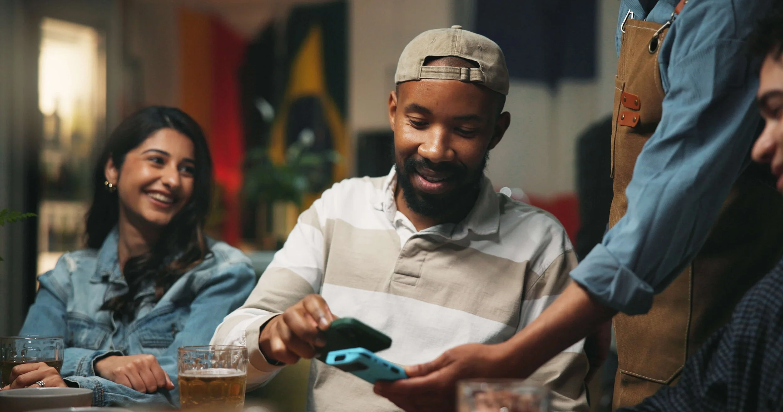 A group of friends socializing at a bar, with one person showing something on their phone to others. The scene appears lively and friendly.