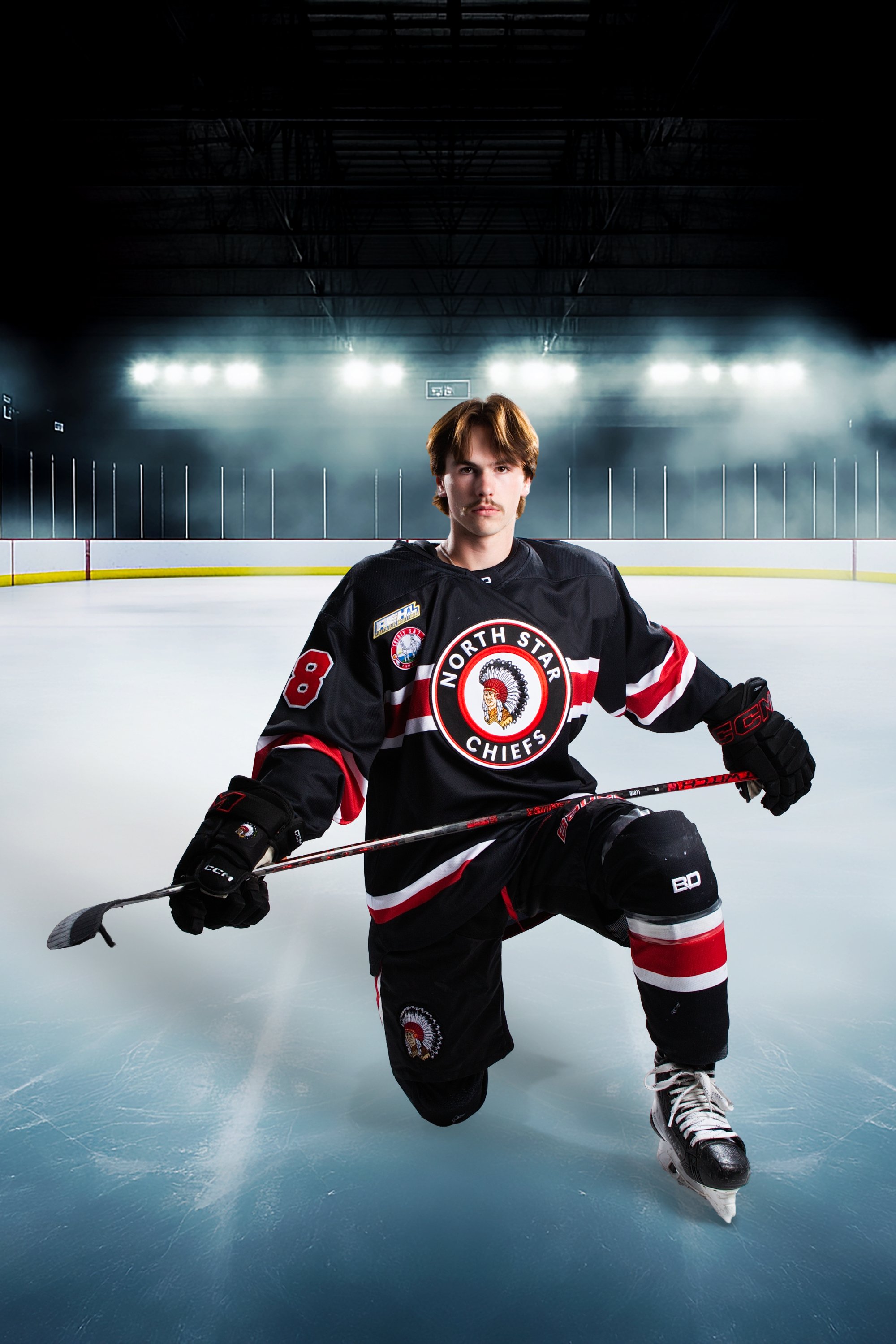 A young male hockey player kneeling on the ice in an indoor hockey rink, wearing a black and red North Star Chiefs jersey, holding a hockey stick, with ice rink boards and bright overhead lights in the background.