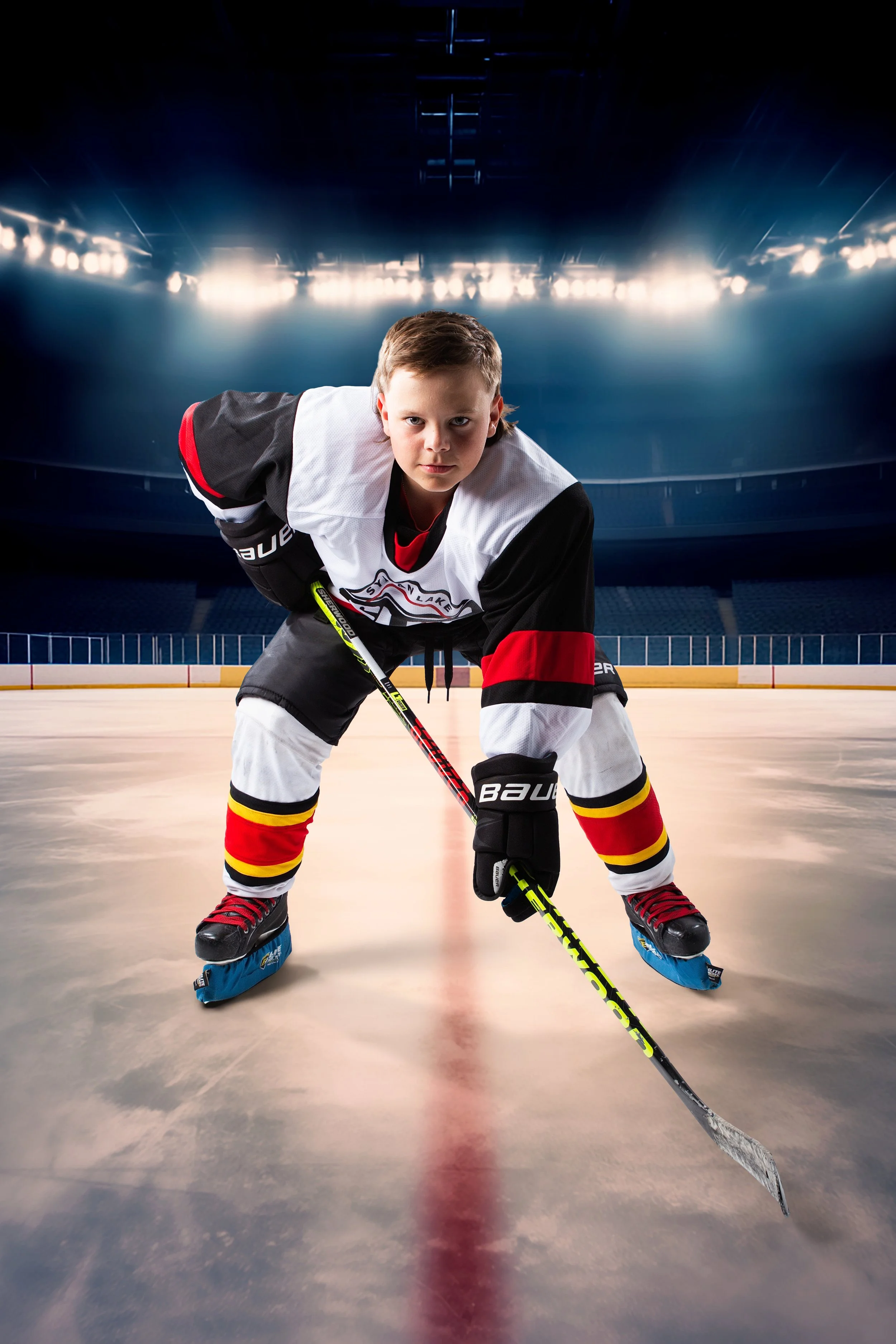 A young boy in ice hockey gear, including skates, gloves, and a jersey, stands on an ice rink inside a large, brightly lit stadium, looking directly at the camera in a ready stance.