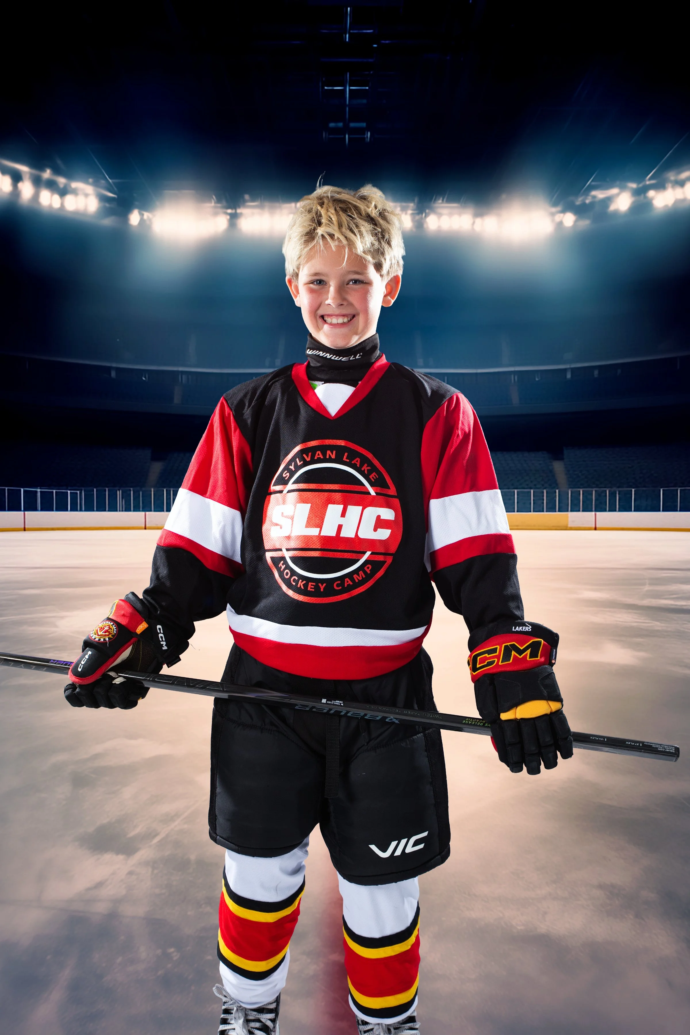 Young boy wearing a red, black, and white hockey jersey and gloves, standing on an ice rink with a smile, holding a hockey stick, in an indoor hockey arena.