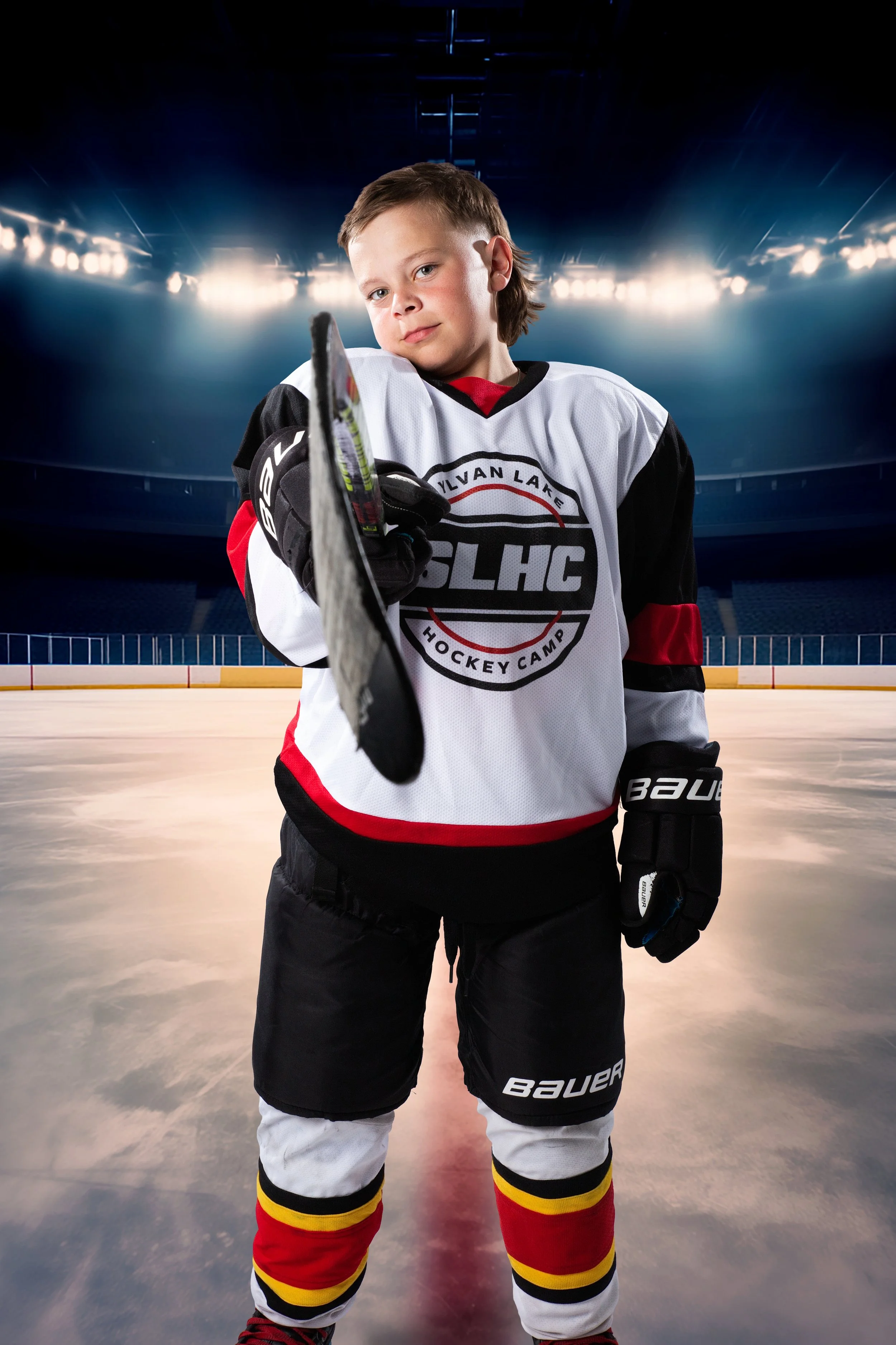 Young hockey player in a black, red, yellow, and white uniform standing on an ice rink with bright arena lights overhead, holding a hockey stick and looking at the camera.