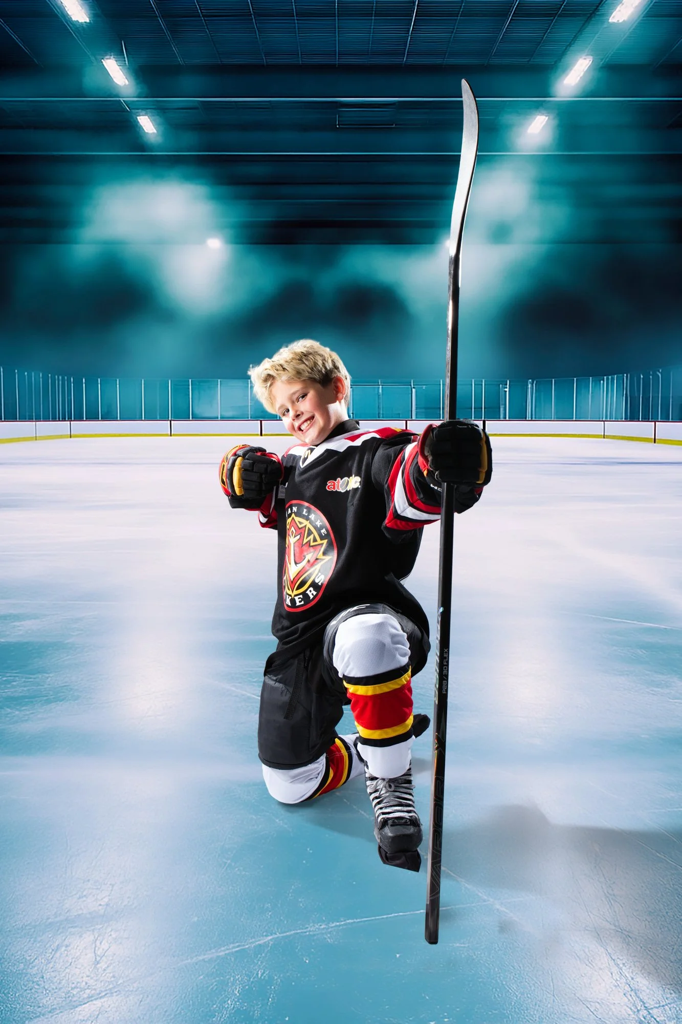 A young boy in hockey gear is kneeling on the ice in an indoor hockey rink, smiling and holding up a hockey stick. The rink is well lit with blue tones.