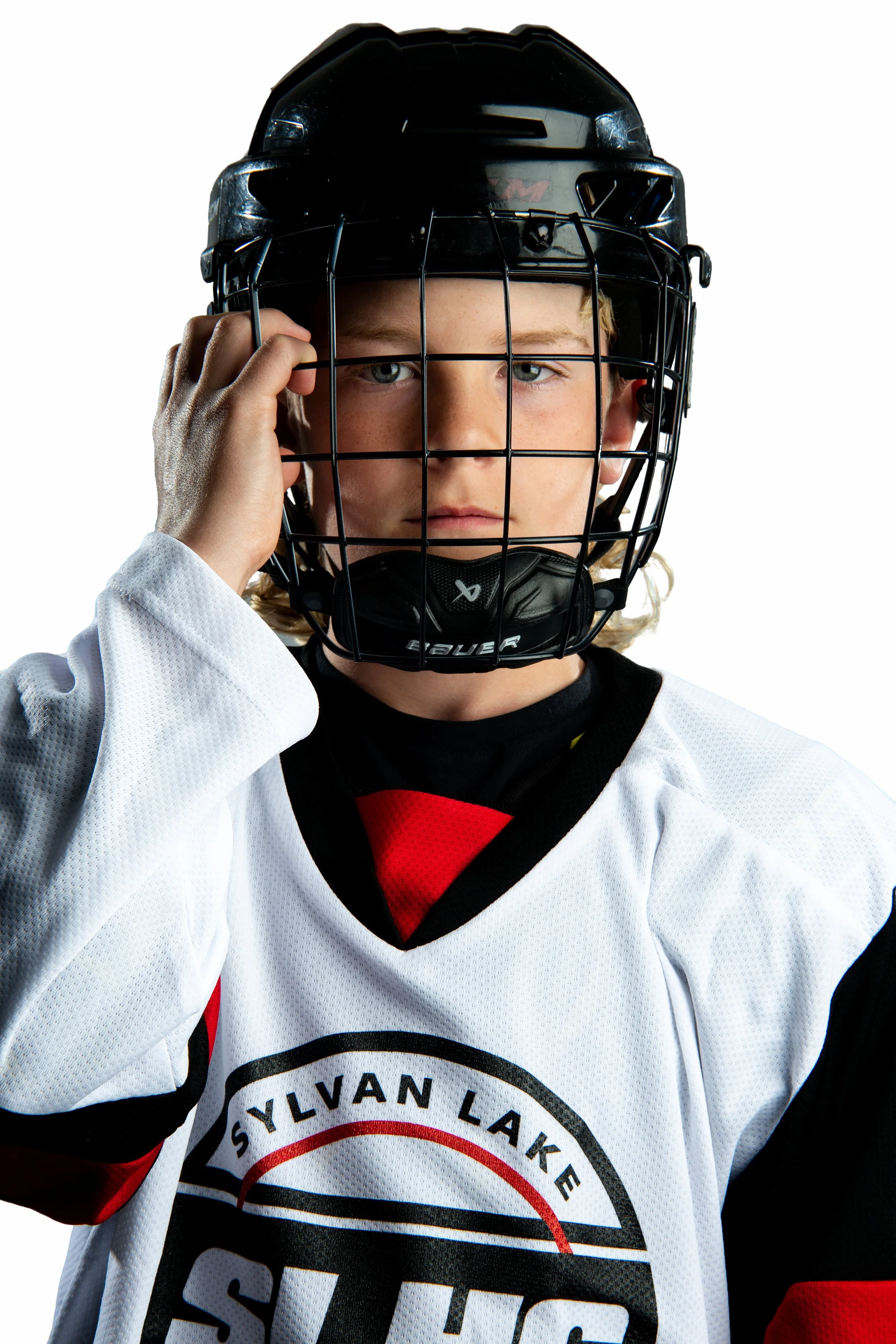 Young hockey player wearing a white jersey with black and red accents, black helmet with face cage, and hockey gloves, holding helmet with one hand, on a white background.