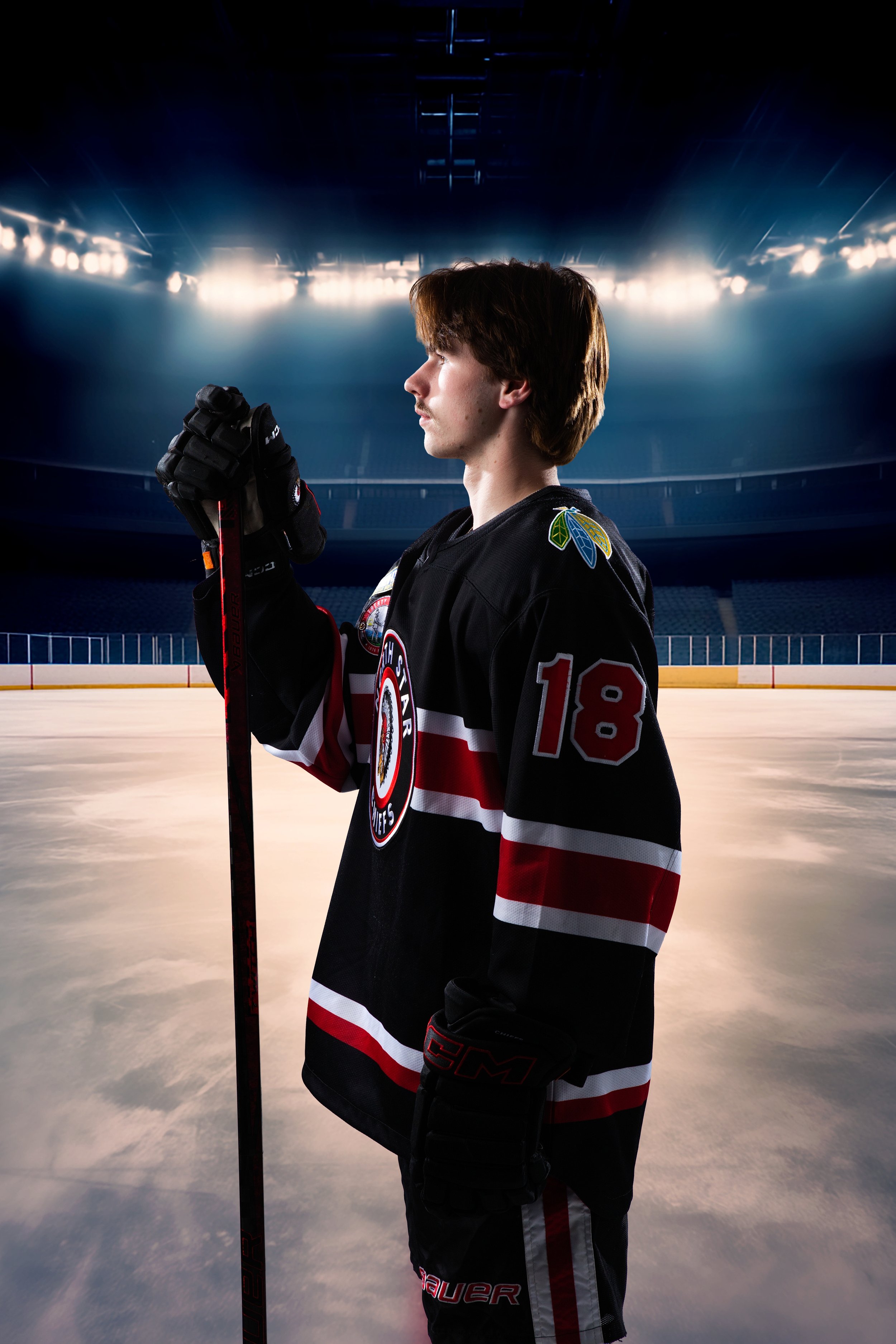 A young male hockey player dressed in black with red and white accents, holding a hockey stick, standing on an ice rink with stadium lights overhead and an empty seating area in the background.