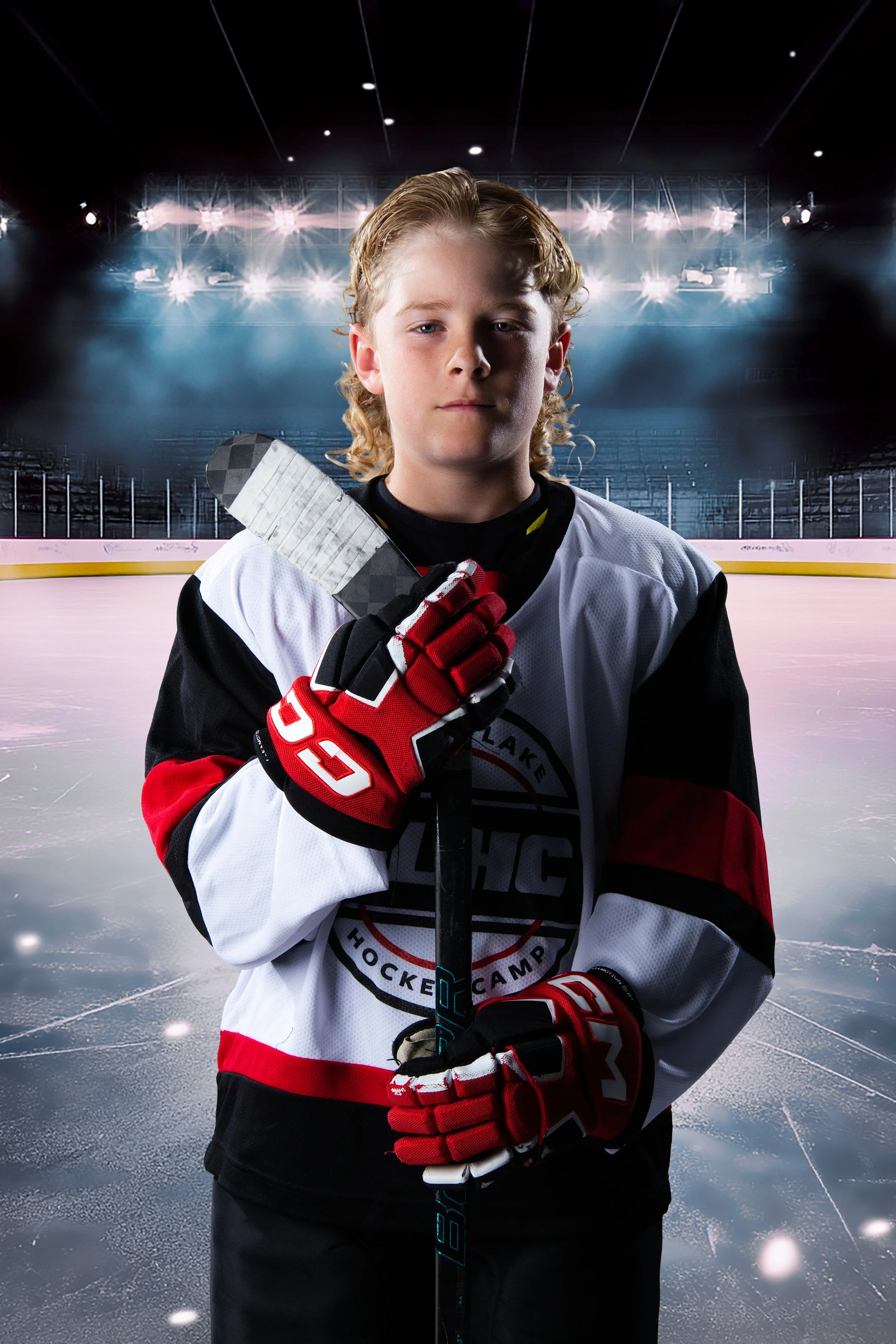 A young male hockey player with long blonde hair stands on an ice rink holding a hockey stick close to his face. He's wearing a white hockey jersey with black, red, and gray accents and red gloves. The background shows bright stadium lights and a dark, empty arena.