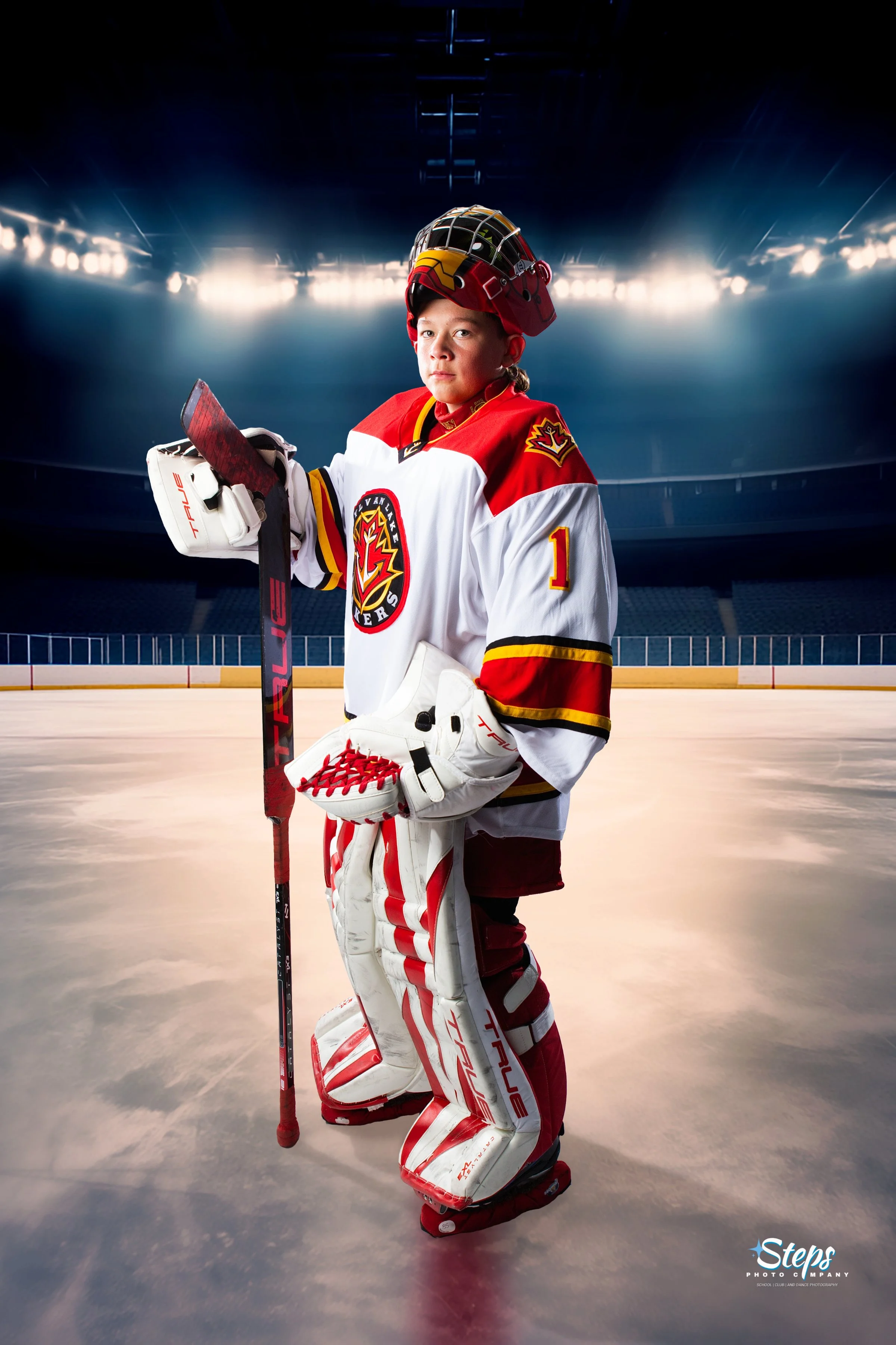 A young hockey player dressed in team gear, including a red, white, and yellow jersey and red and white skates, stands on the ice rink holding a hockey stick. The background features an empty ice rink with bright lights and a dark, indoor arena.