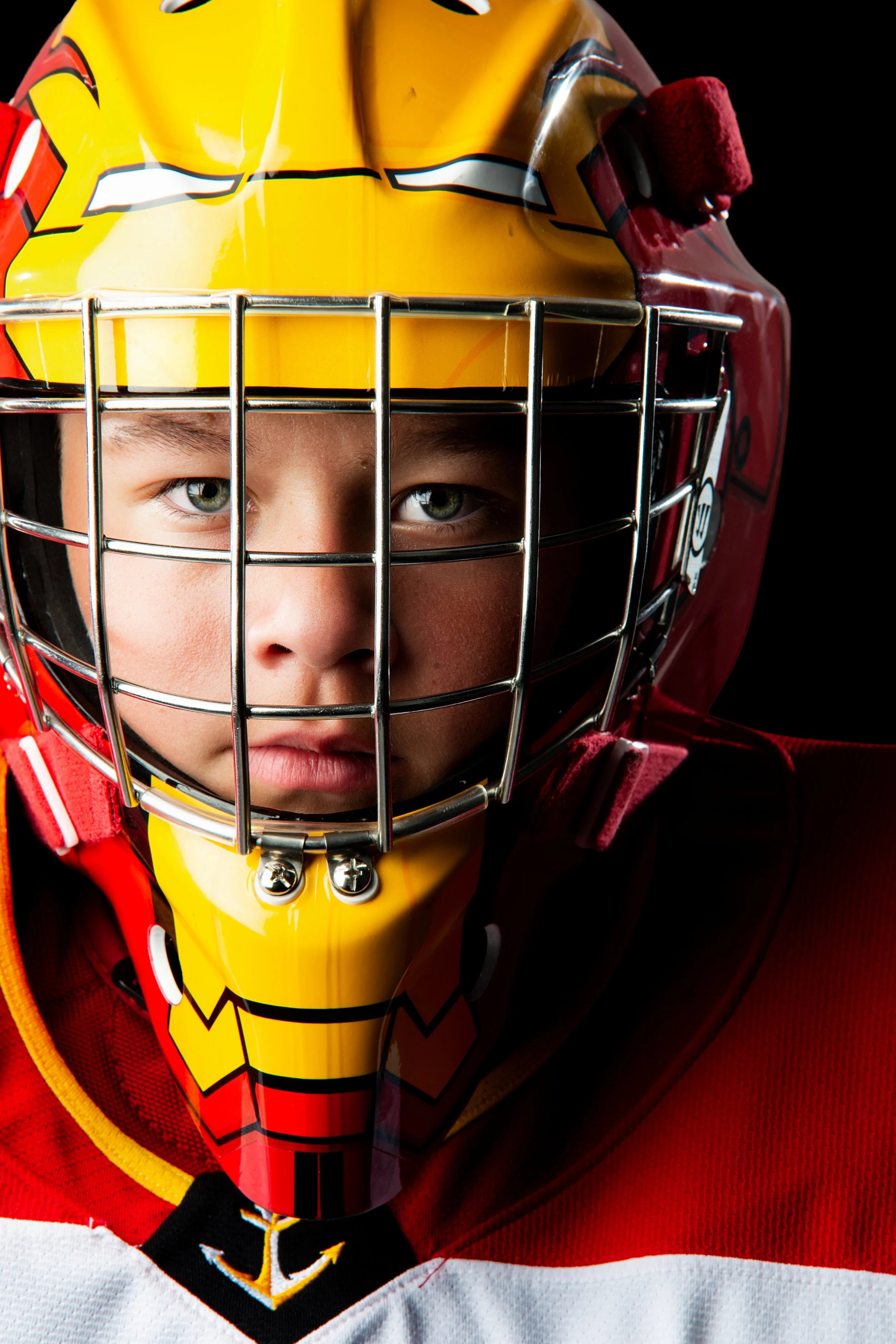 Close-up of a young hockey player wearing a yellow helmet with a red and black logo, a face cage, and red gear, against a black background.