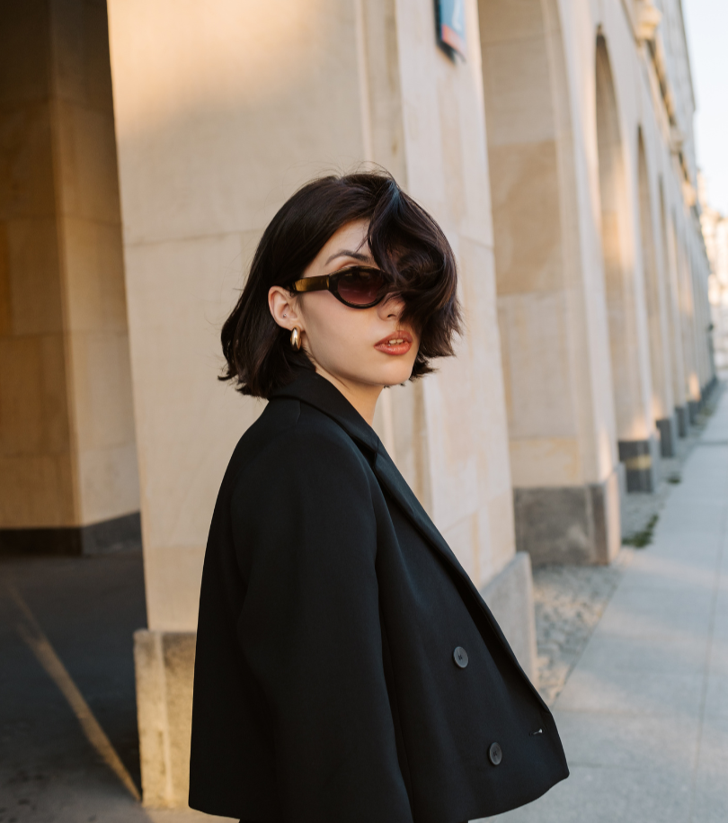A young woman with short dark hair wearing large sunglasses, gold hoop earrings, a black blazer, and a black top, standing on a city sidewalk with stone building walls in the background.