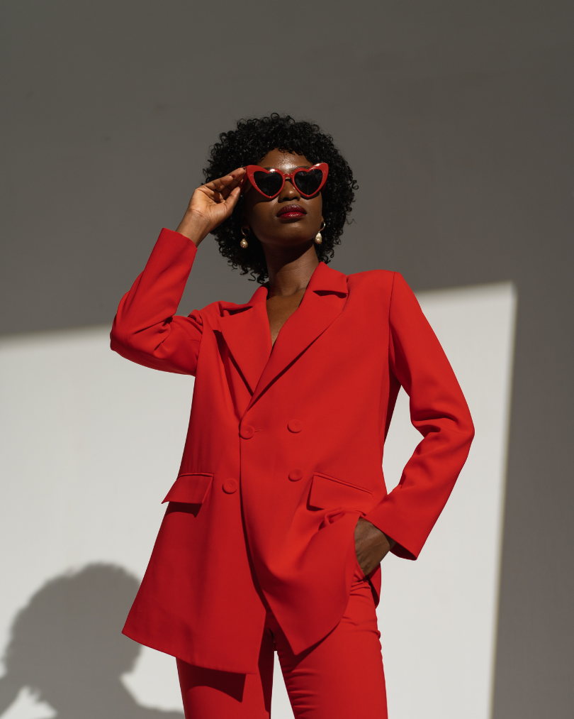 A woman wearing a red suit and heart-shaped sunglasses, standing against a plain background with a shadow cast on the wall.