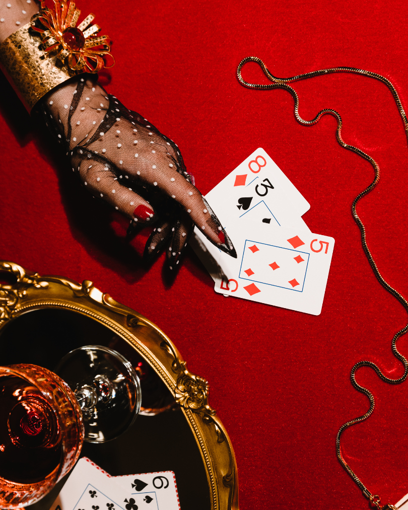 A woman wearing red nail polish and black fishnet gloves with white polka dots, holding a card from a deck of playing cards on a red velvet surface. There are four playing cards visible, including a five, eight, and three, along with a diamond card, and a quarter of a gold ornate mirror with a glass of red wine is also present. A necklace with small black beads is draped on the surface.