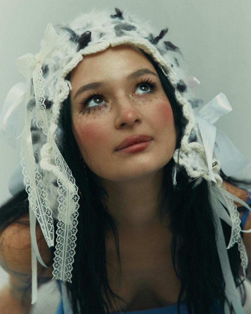A young woman with dark hair, blue eyes, and freckles looking upwards, wearing a white, lacy bonnet with ribbons and crochet details