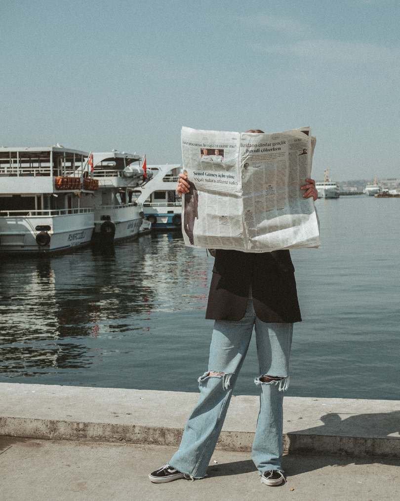 Person standing at a waterfront, holding a large newspaper in front of their face, with boats docked in the background.