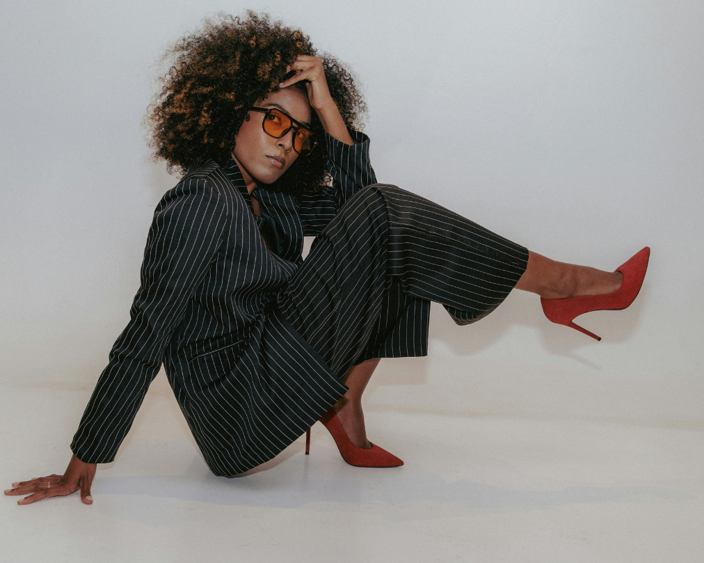Woman with curly hair wearing a black pinstripe suit, red high heels, and orange-tinted glasses, sitting on the floor with one leg raised and touching her forehead, against a plain white background.