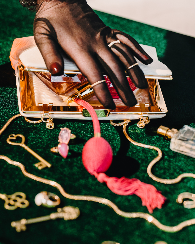 A hand wearing a black sheer glove with rings is reaching into an open, shiny gold clutch purse. The purse contains a white notebook or compact mirror. On a green surface, various jewelry pieces including gold chains, keys, rings, earrings, and a pink pouch connected to a chain are displayed, with a perfume bottle visible in the corner.