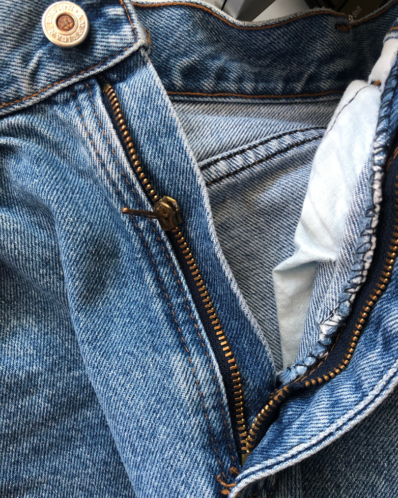 Close-up of a partially unzipped pair of blue denim jeans showing the zipper and button with stitching details.