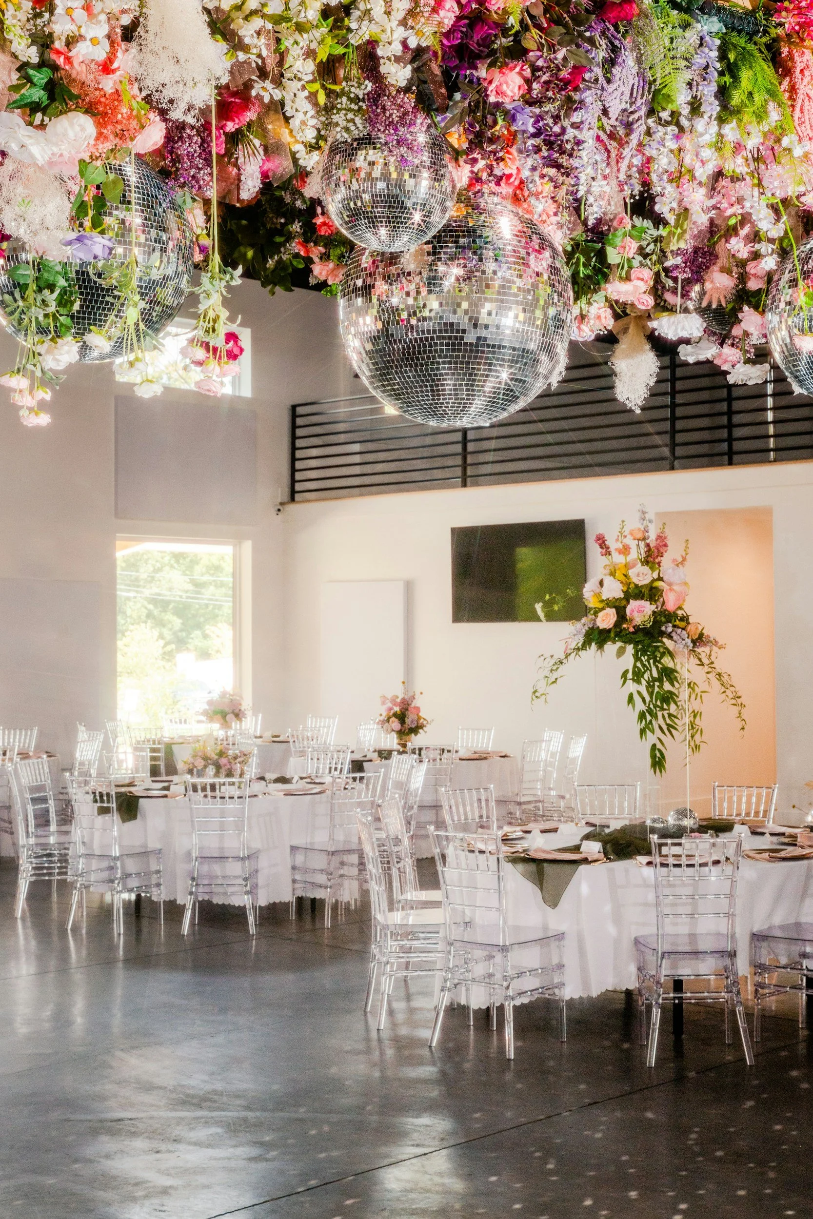 Elegant event space with round tables covered in white tablecloths, decorated with floral centerpieces, clear acrylic chairs, and hanging disco balls amid colorful flowers on the ceiling.