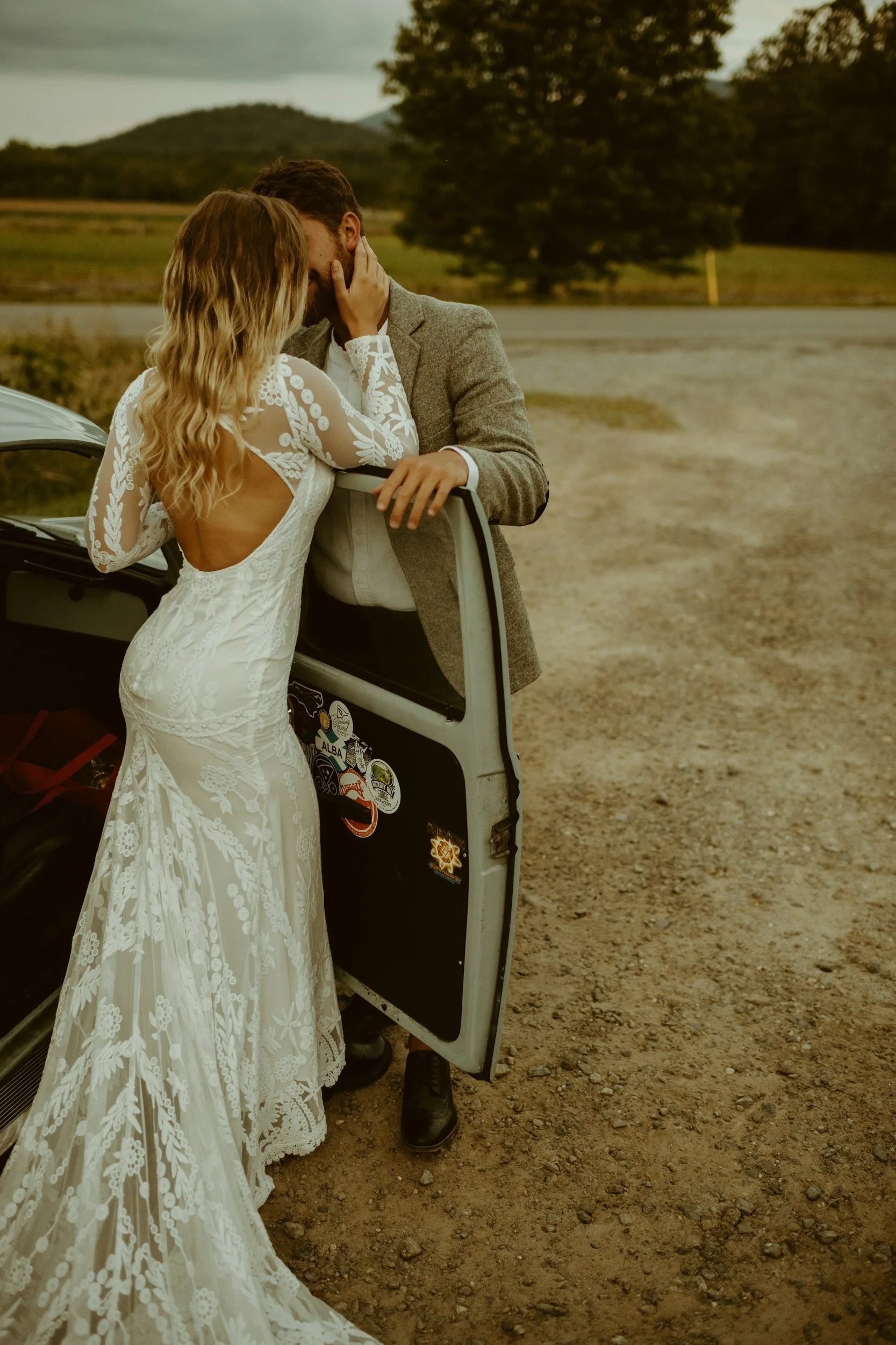 A bride and groom sharing a kiss outside near a car with stickers, with a scenic landscape in the background.