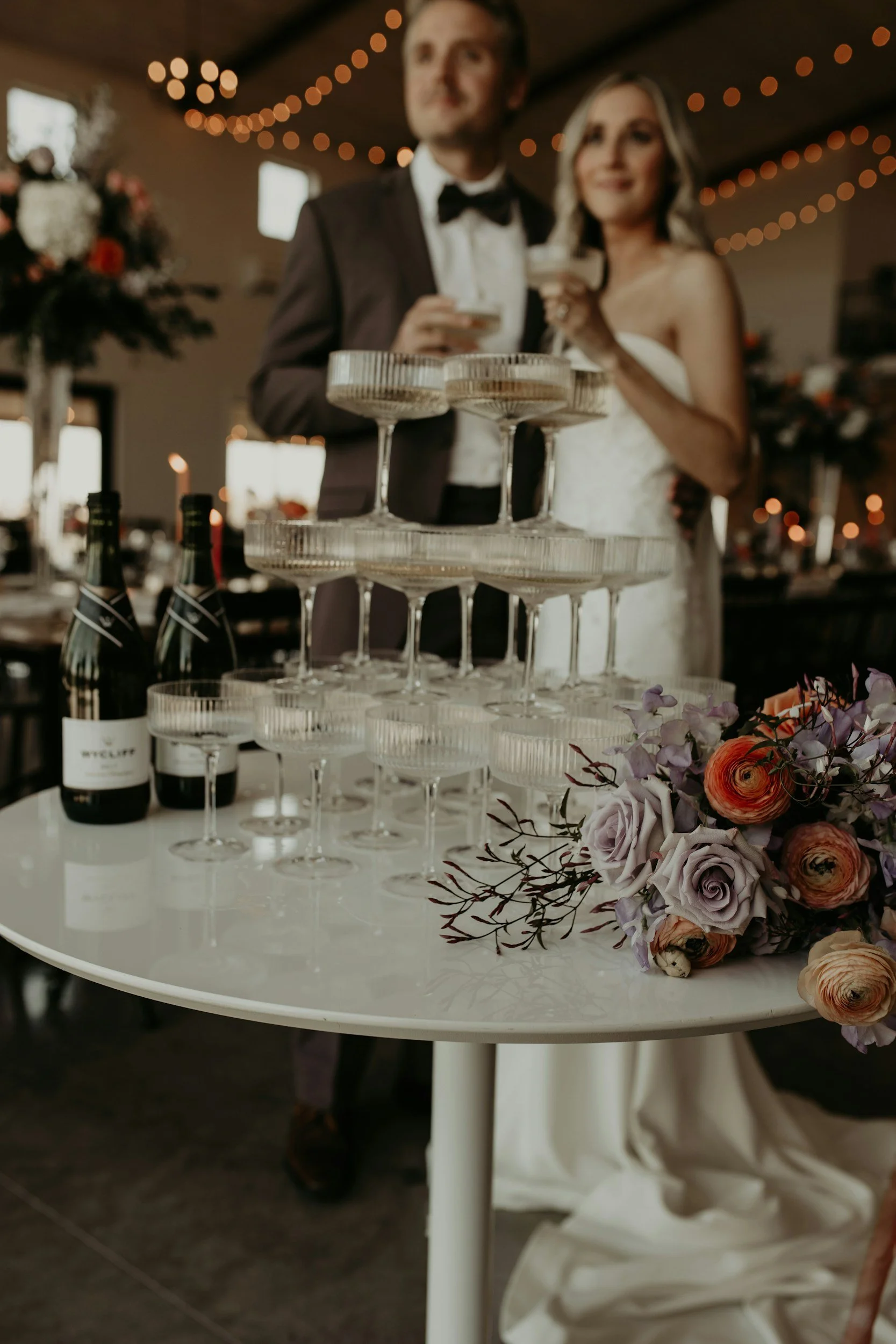 Wedding celebration with a champagne tower, bottles of champagne, and a floral centerpiece on a white table, with a bride and groom in the background.