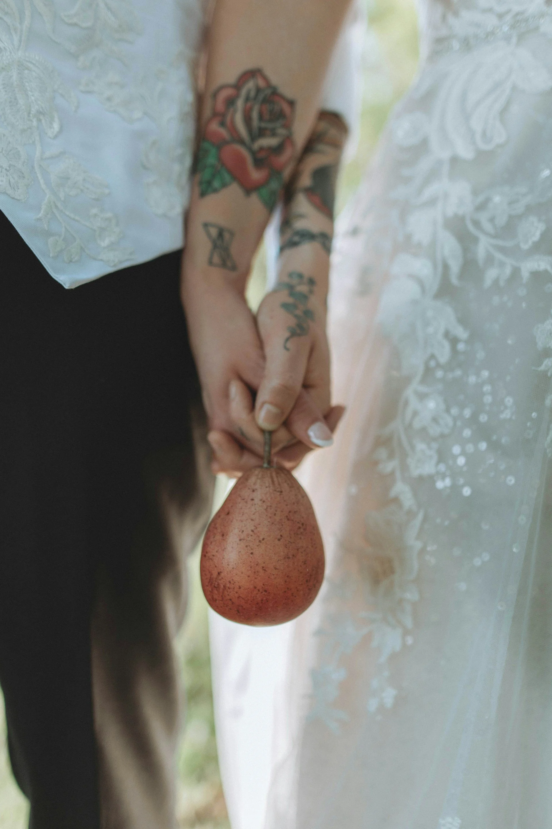 Close-up of intertwined hands holding a pear-shaped object, with visible tattoos on the arms, against a background of a lace curtain.