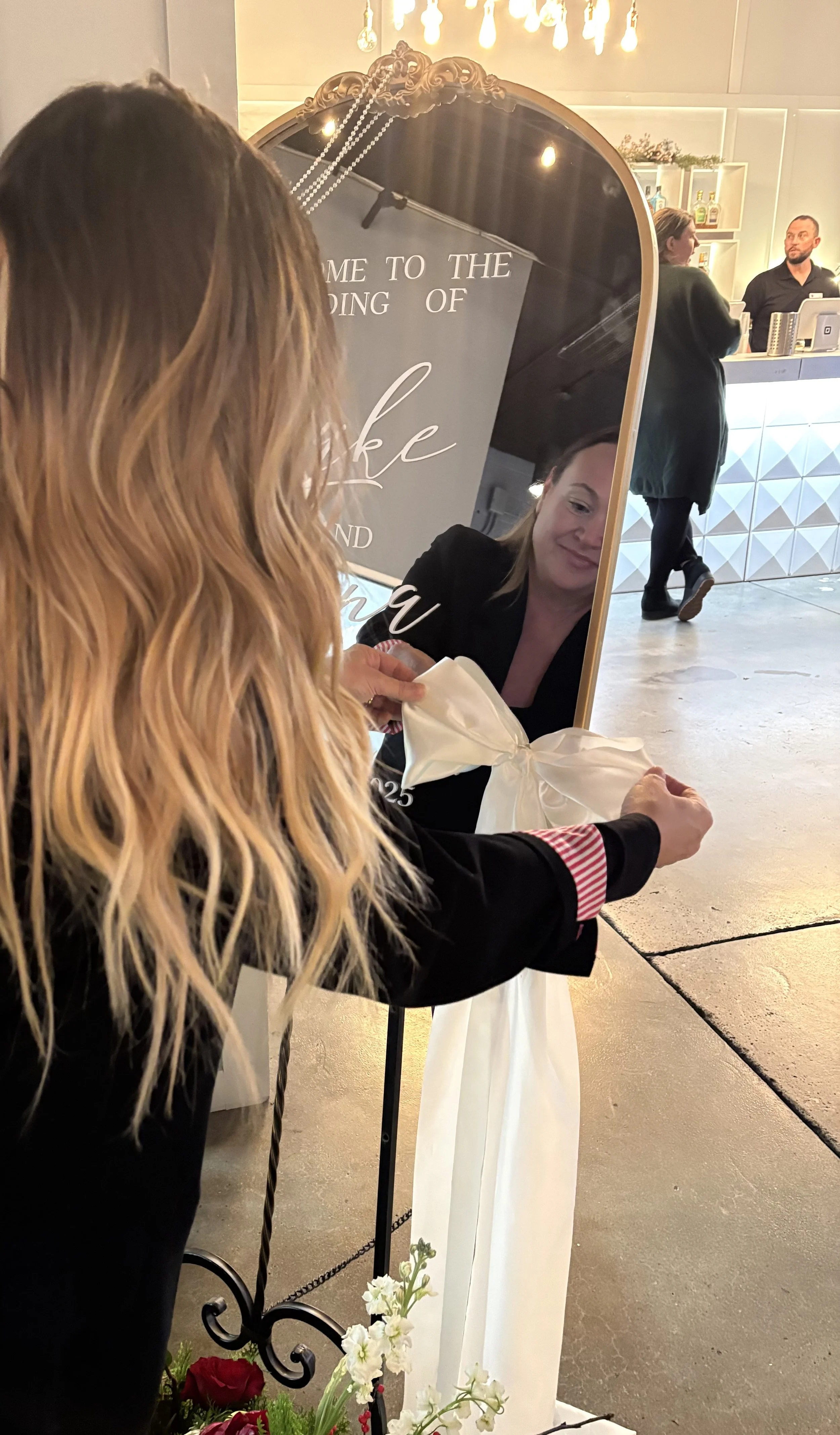 A woman is holding a white ribbon with a bow, tying it in front of a large mirror. Behind her, a woman with blonde hair and a woman with short hair are visible in the reflection. The setting appears to be a celebration, possibly a grand opening or special event, with a decorated sign in the background and a modern reception desk with two people talking.