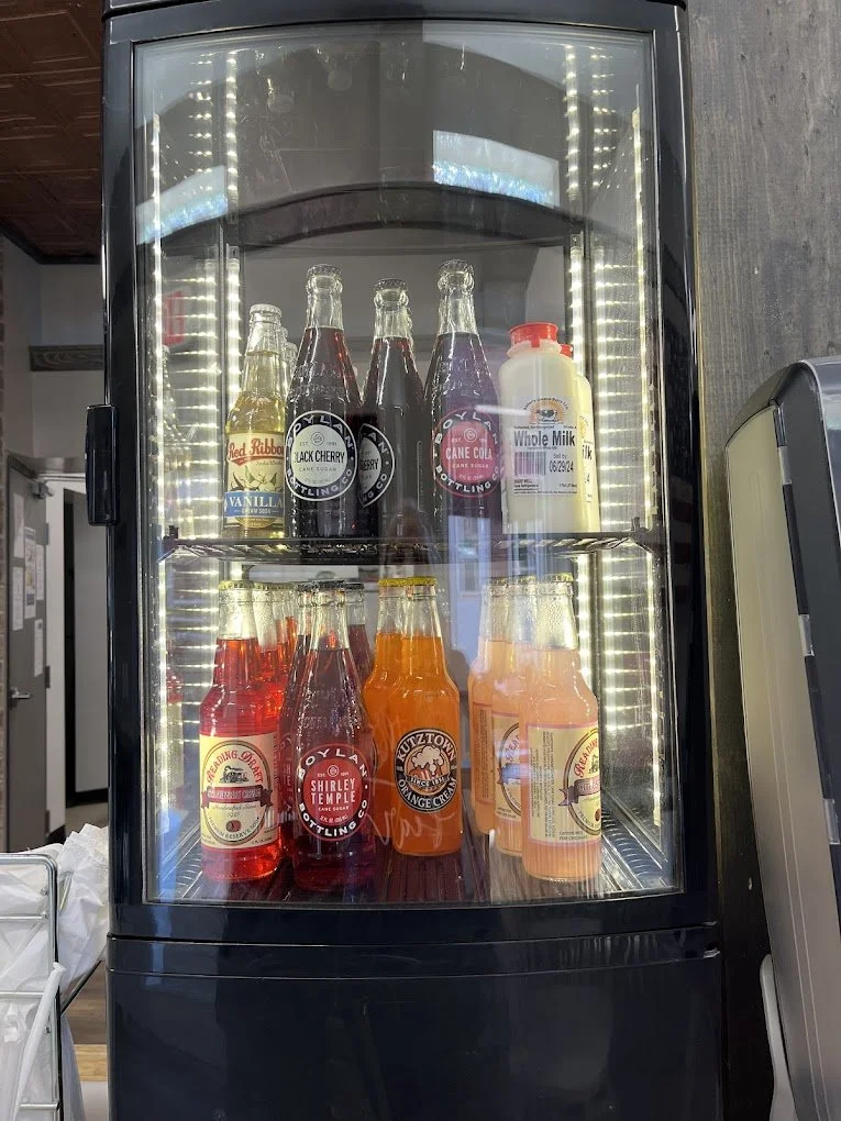 A refrigerator filled with various bottles of soda and milk, including red ribbon, black cherry, cane cola, whole milk, and other flavored sodas in glass bottles, behind a glass door with LED lighting.