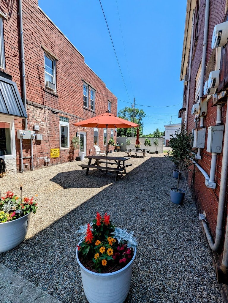 Outdoor courtyard with gravel ground, potted flowers, picnic tables, umbrellas, potted trees, against red brick building walls, with clear blue sky.