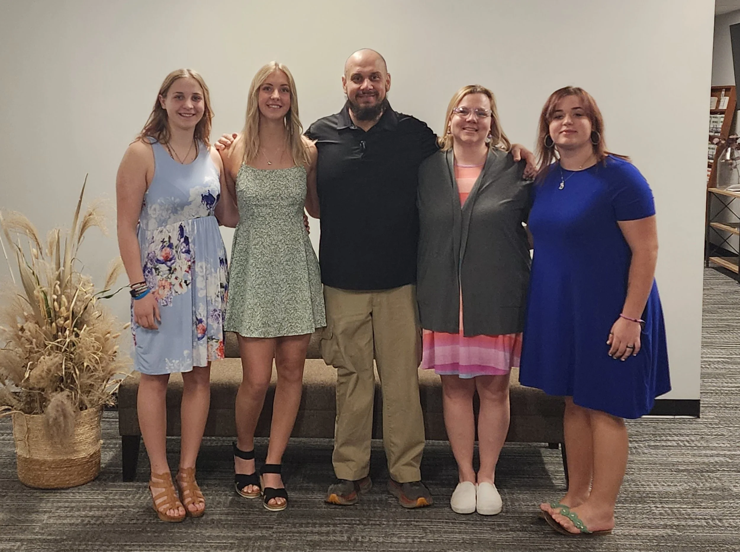 Group of five people standing together indoors, smiling, with a beige plant to the left and a neutral background.