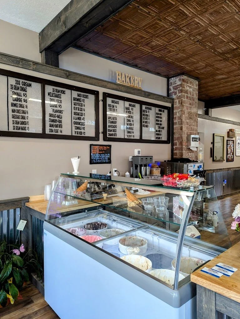 Ice cream display freezer with various ice cream flavors in a bakery shop, with menu boards overhead listing baked goods and flavors.