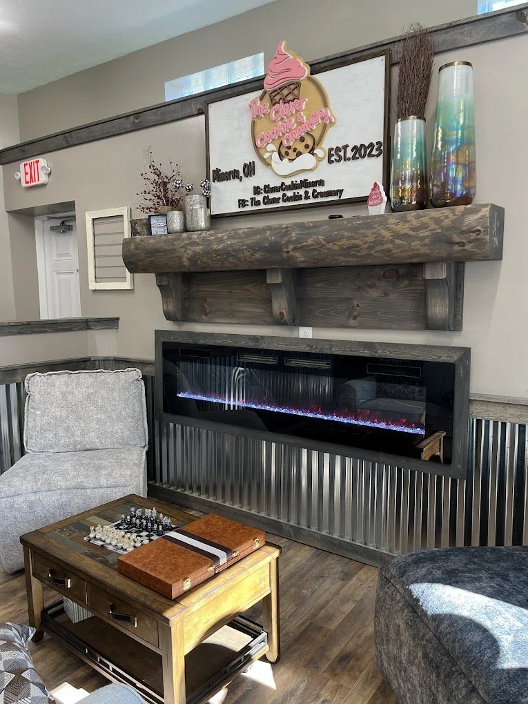 Living room with a fireplace, gray armchair, wooden coffee table with chess and books, decorative vases, and a sign reading 'The Clever Cookie & Bakery, Memory, OH, EST. 2023' with ice cream cone and cookie graphics.
