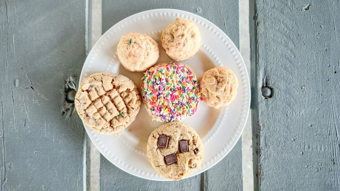 A white plate with six cookies of different flavors, including a sprinkled cookie, a chocolate chip cookie, a peanut butter cookie, a sugar cookie with a criss-cross pattern, and two coconut macaroons, placed on a rustic wooden table.