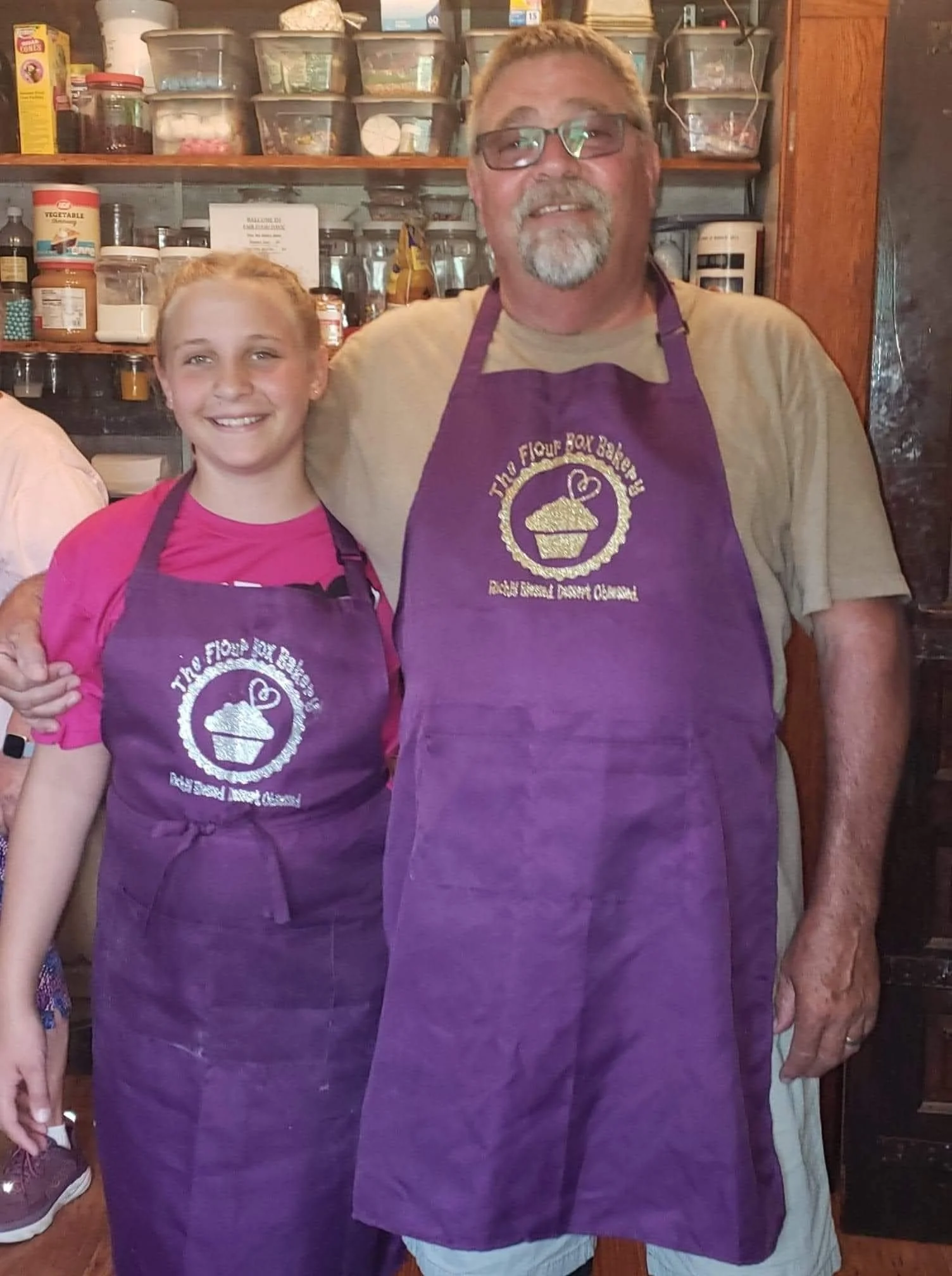 A smiling young girl and an older man wearing purple aprons with a cupcake and heart logo, standing in a kitchen with shelves of ingredients behind them.